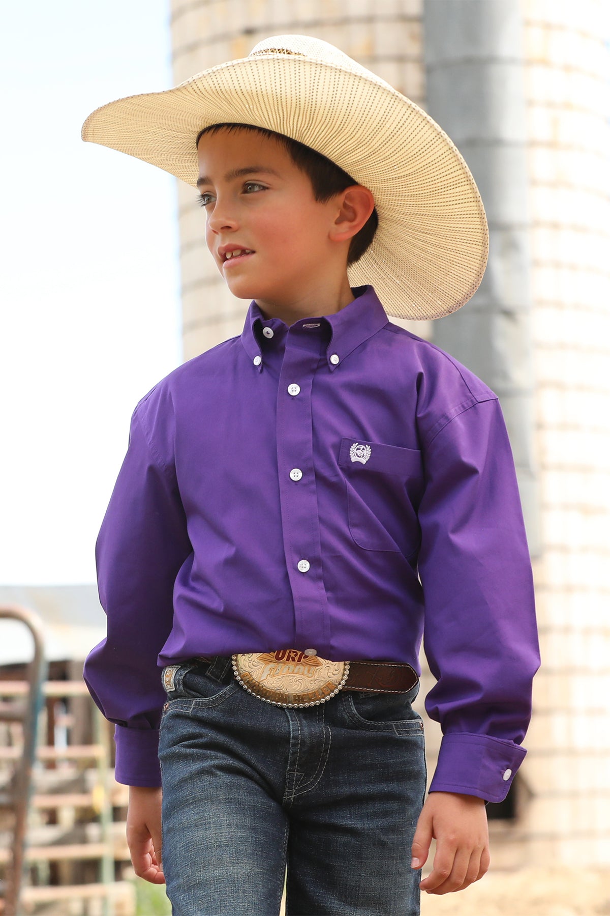 Boy standing outside in front of a silo wearing a purple long sleeve button-down with jeans, a belt and a cowboy hat