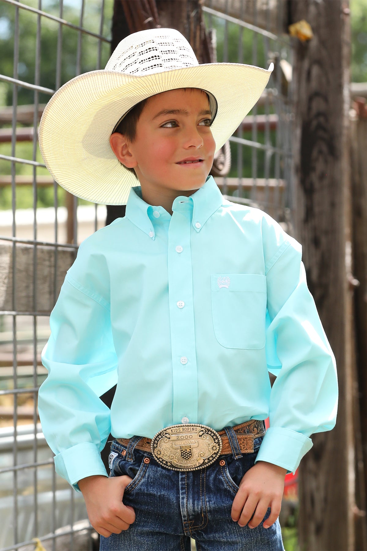 Boy standing outside in front of a fence wearing a long sleeve button-down with jeans, a belt and a cowboy hat