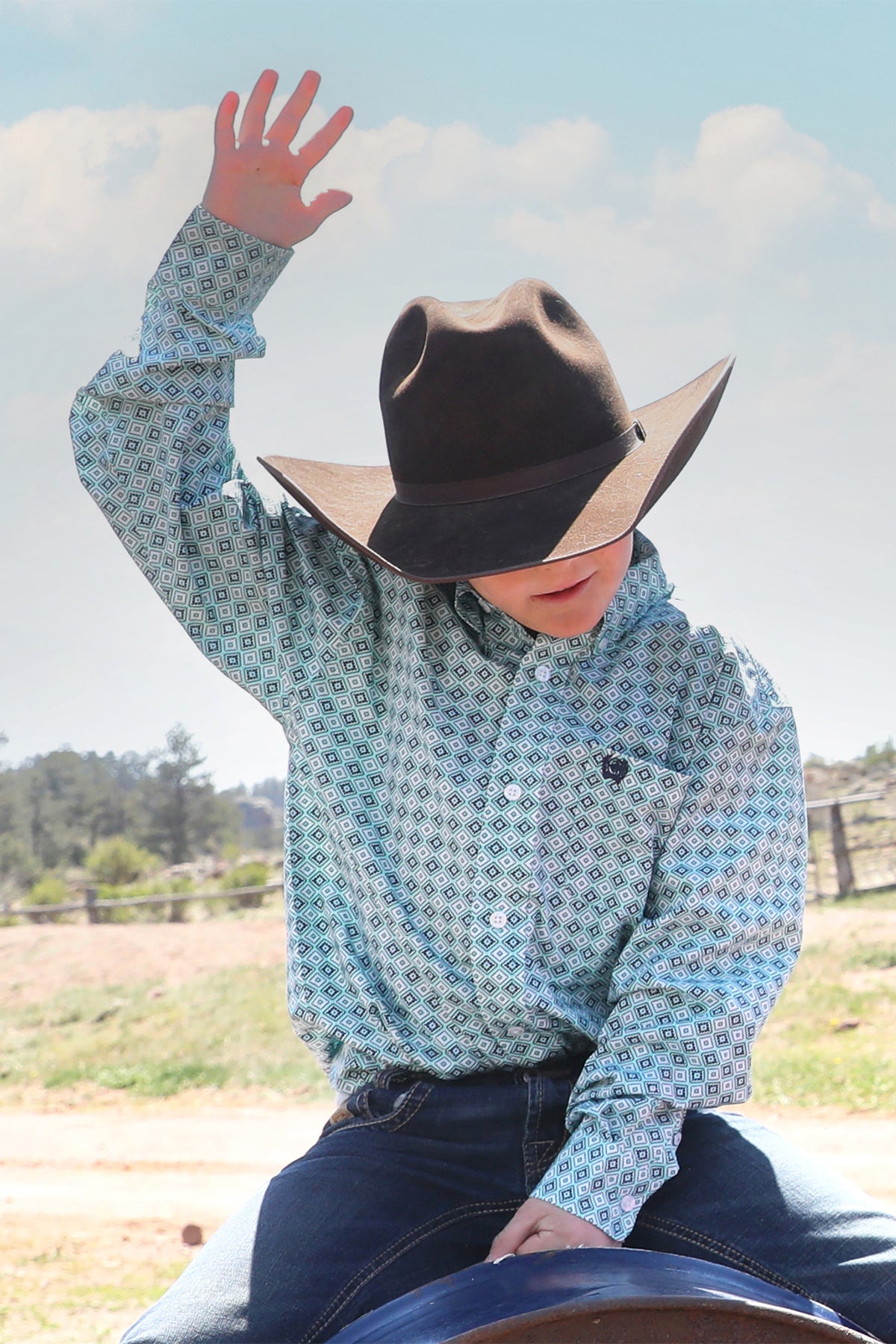Boy wearing a patterned, teal, long sleeve button-down, with jeans and a cowboy hat with his hand in the air