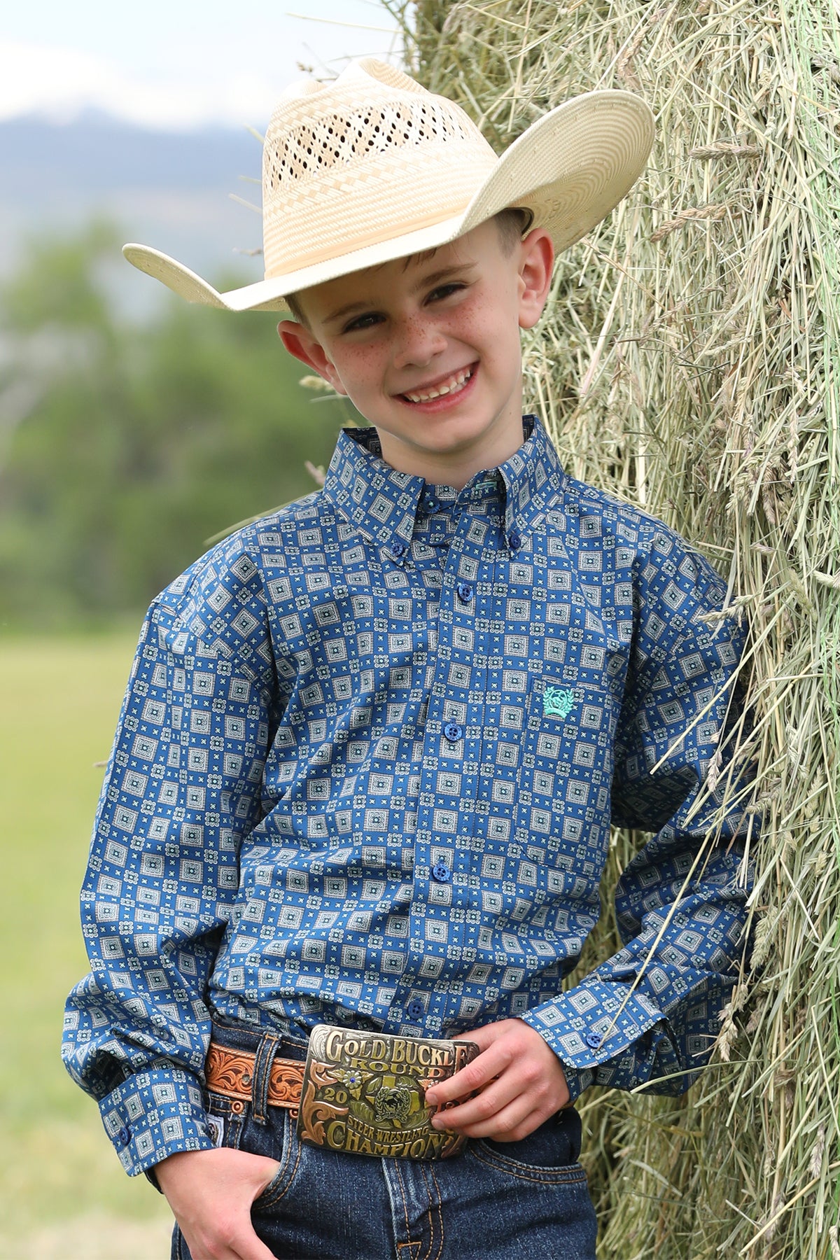 Boy standing outside in front of a bale of hay wearing a patterned long sleeve button-down shirt with jeans, a belt, and a cowboy hat