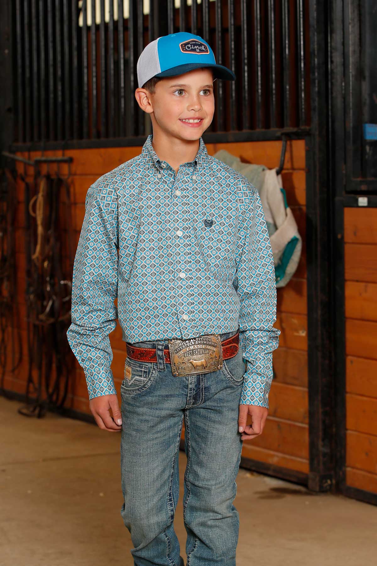Boy standing in a barn, wearing a blue long sleeve button-down, jeans, a western belt, and a hat