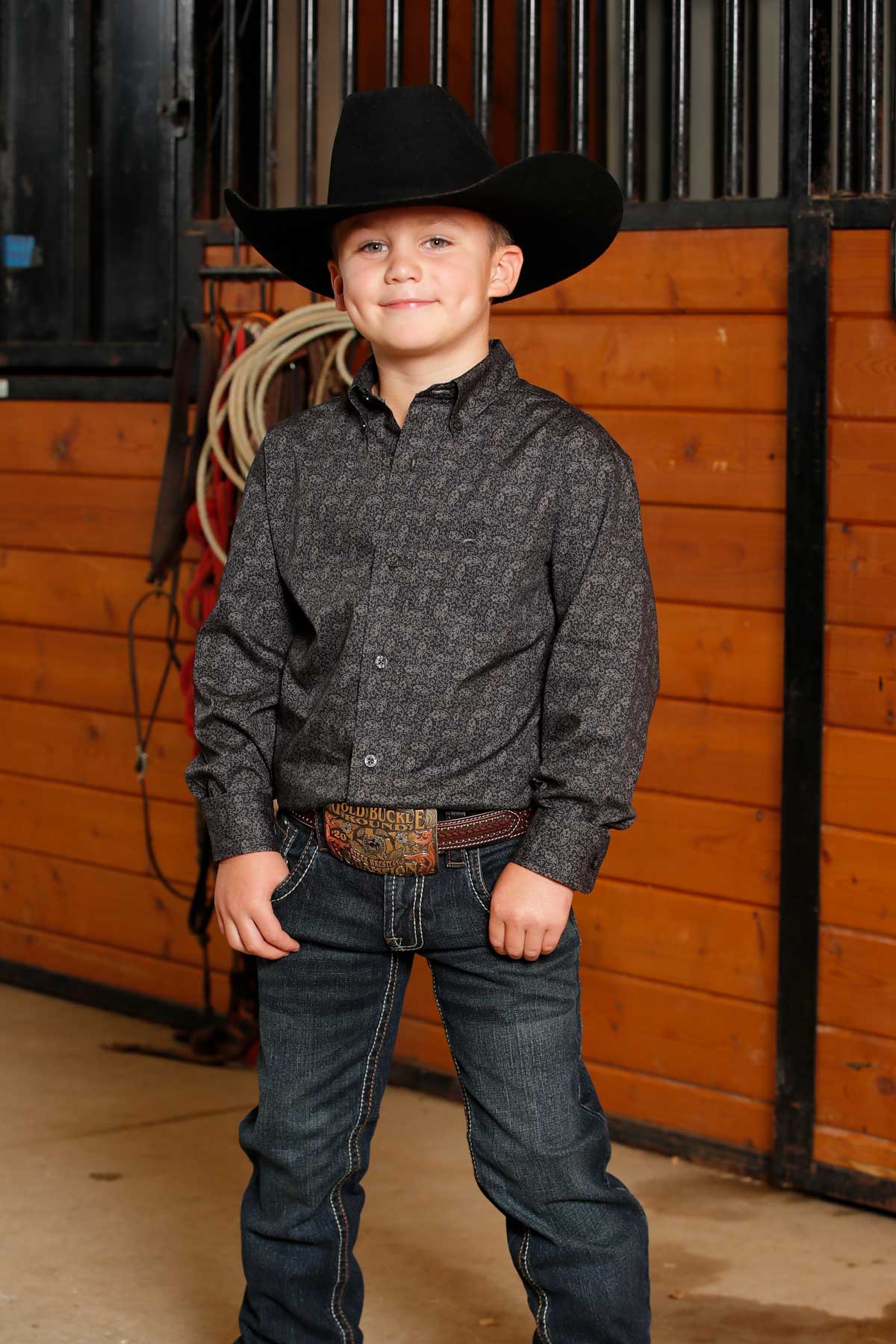 Young boy standing in a barn wearing a gray long sleeve button-down with jeans and a cowboy hat