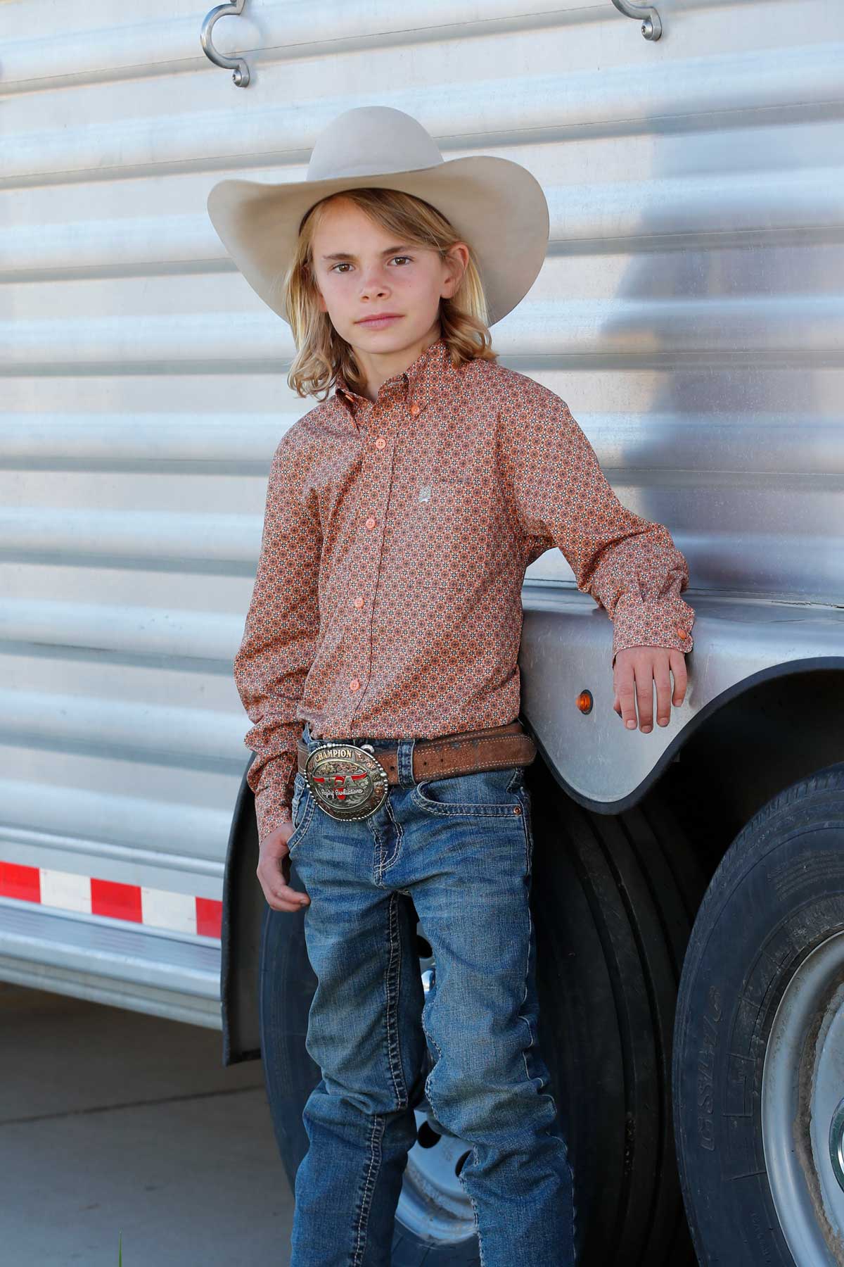 Young boy standing in front of a trailer wearing a coral long sleeve button-down shirt with jeans and a cowboy hat