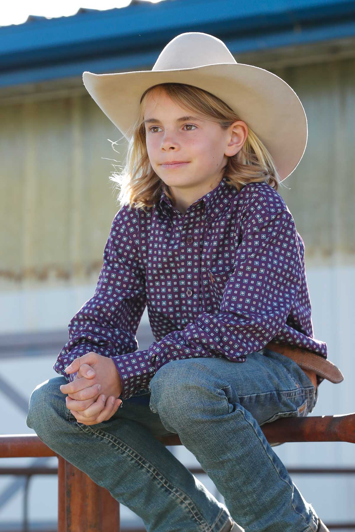 Boy sitting on a fence wearing a purple long sleeve button-down, jeans and a cowboy hat