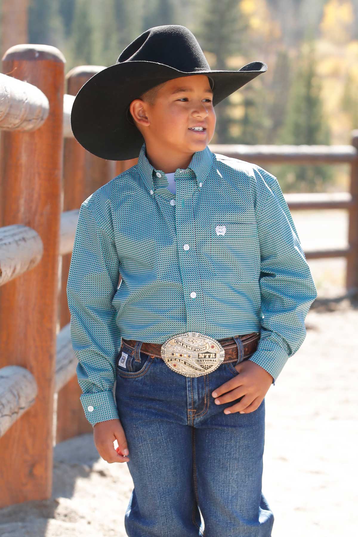 Young boy wearing a teal long sleeve button-down with jeans and a cowboy hat standing in front of a wooden fence