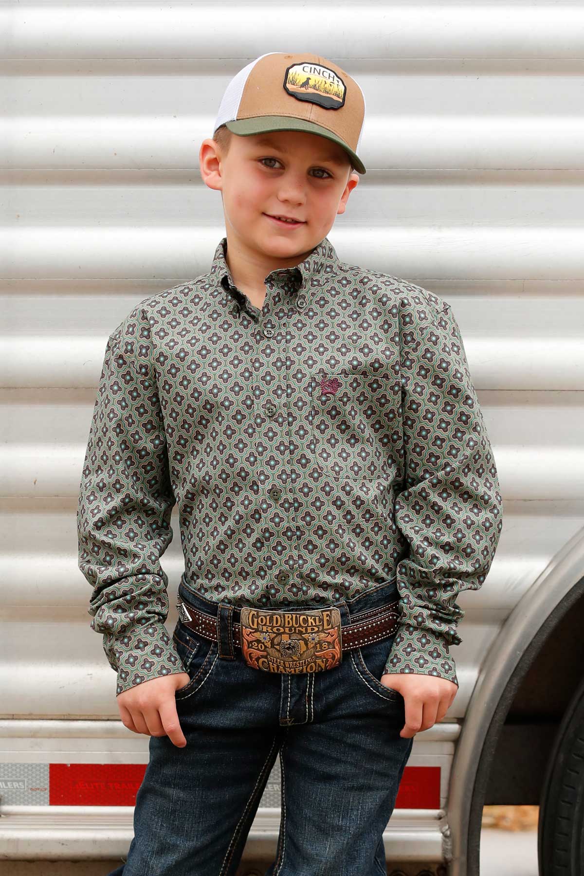 Young boy standing in front of a trailer wearing a green long sleeve button-down with jeans and a ball cap
