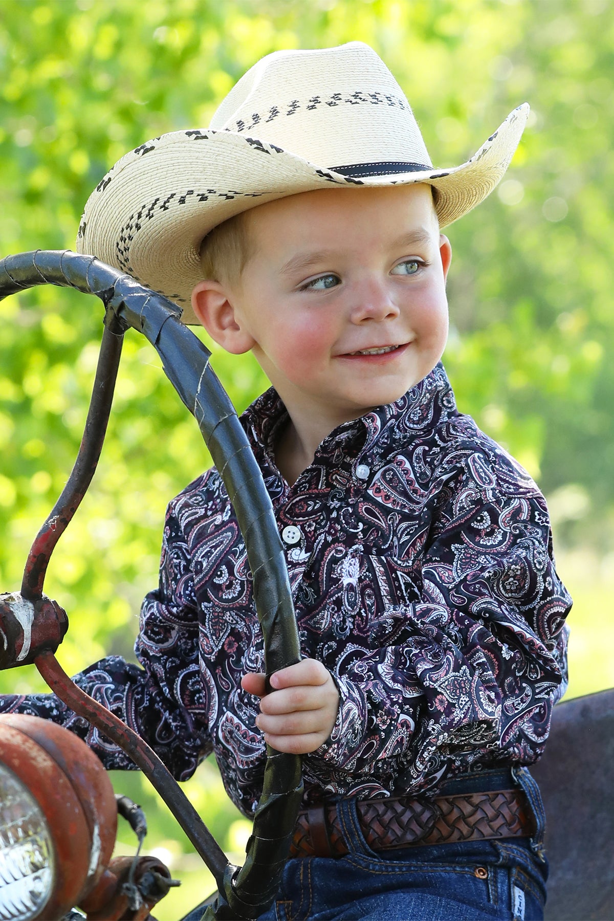 Little boy sitting outside wearing a patterned long sleeve button-down with jeans, a belt, and a cowboy hat