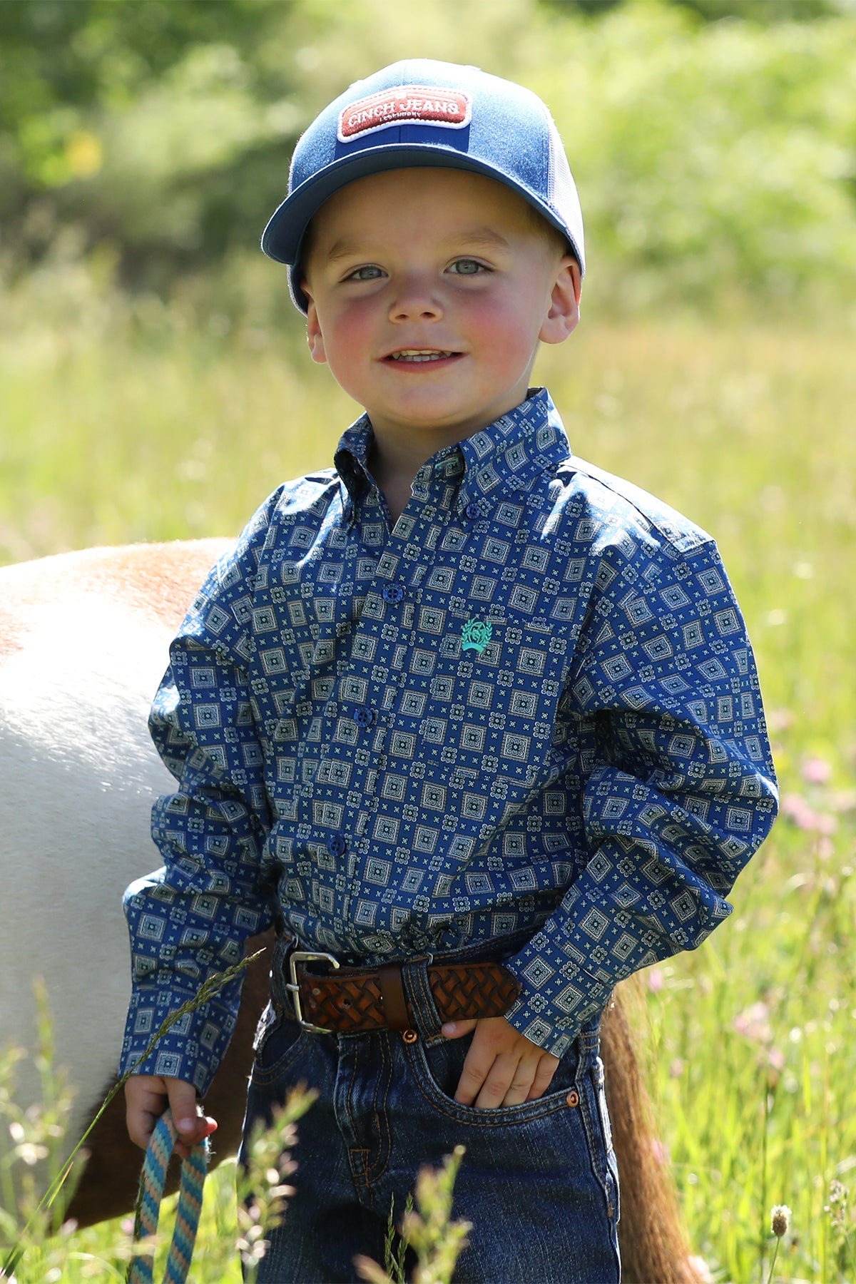 Little boy standing outside in front of a pony wearing a long sleeve button-down with jeans, a belt and a hat