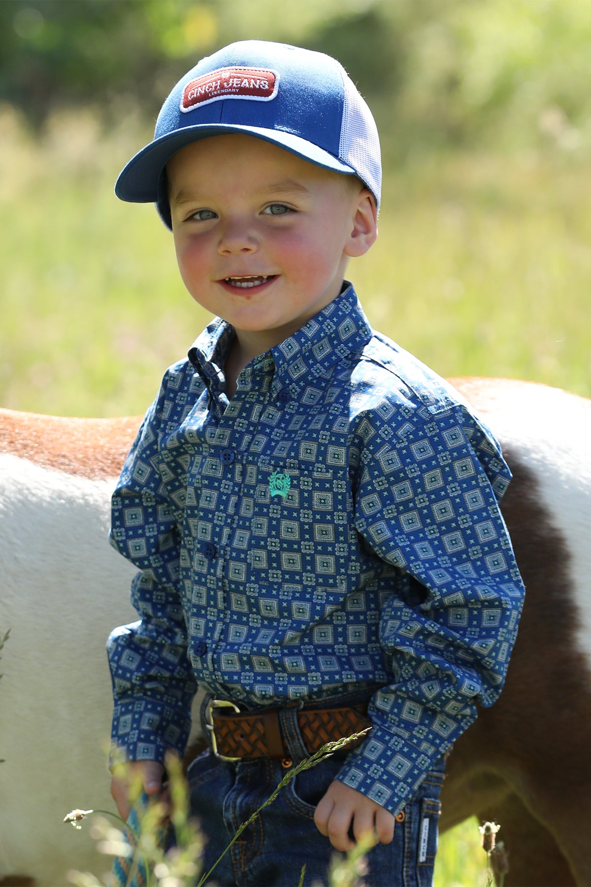 Little boy standing outside in front of a pony wearing a long sleeve button-down with jeans, a belt and a hat