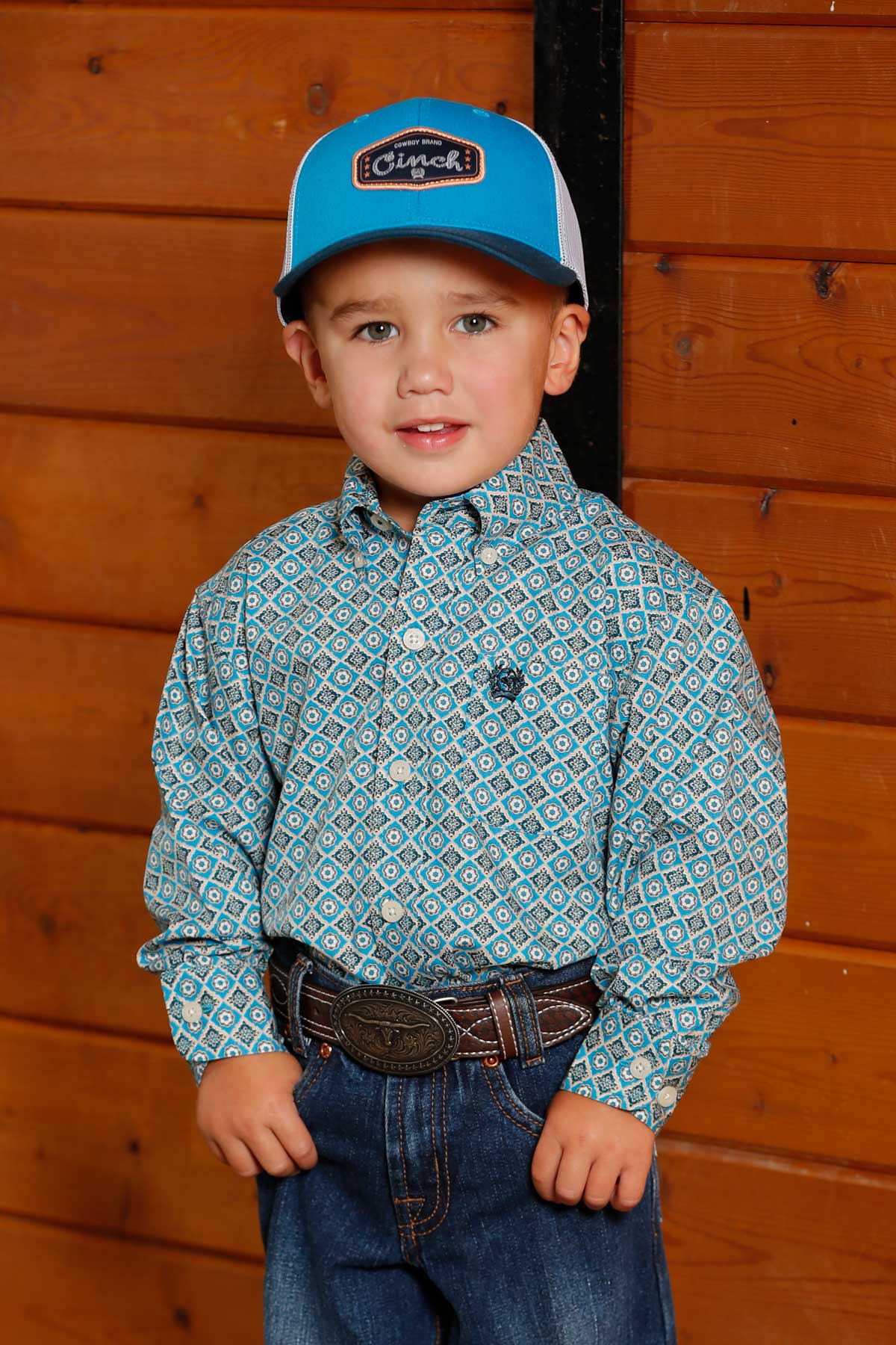 Little boy standing wearing a long sleeve button-down and jeans with a ball cap