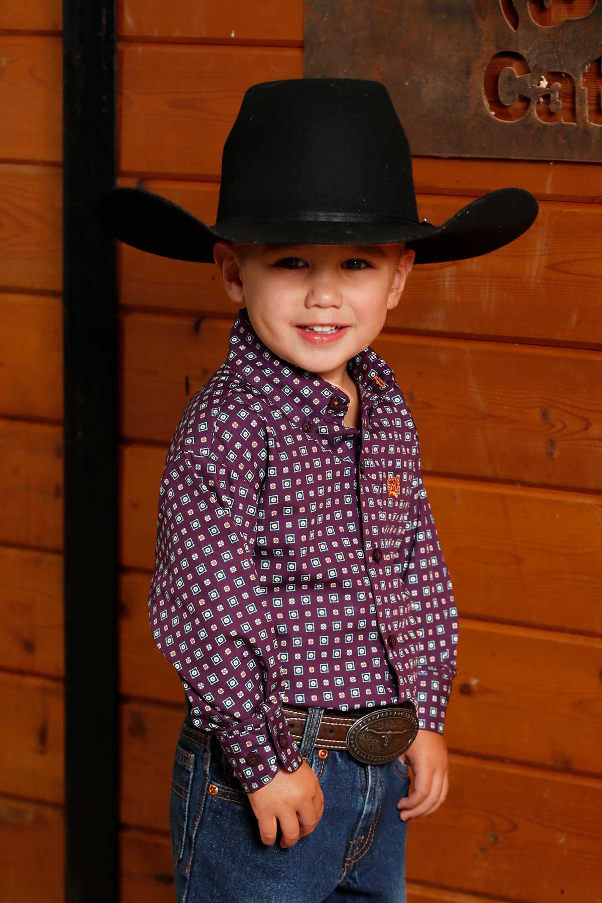 Young boy wearing a purple long sleeve button-down with jeans and a cowboy hat in front of a wood background
