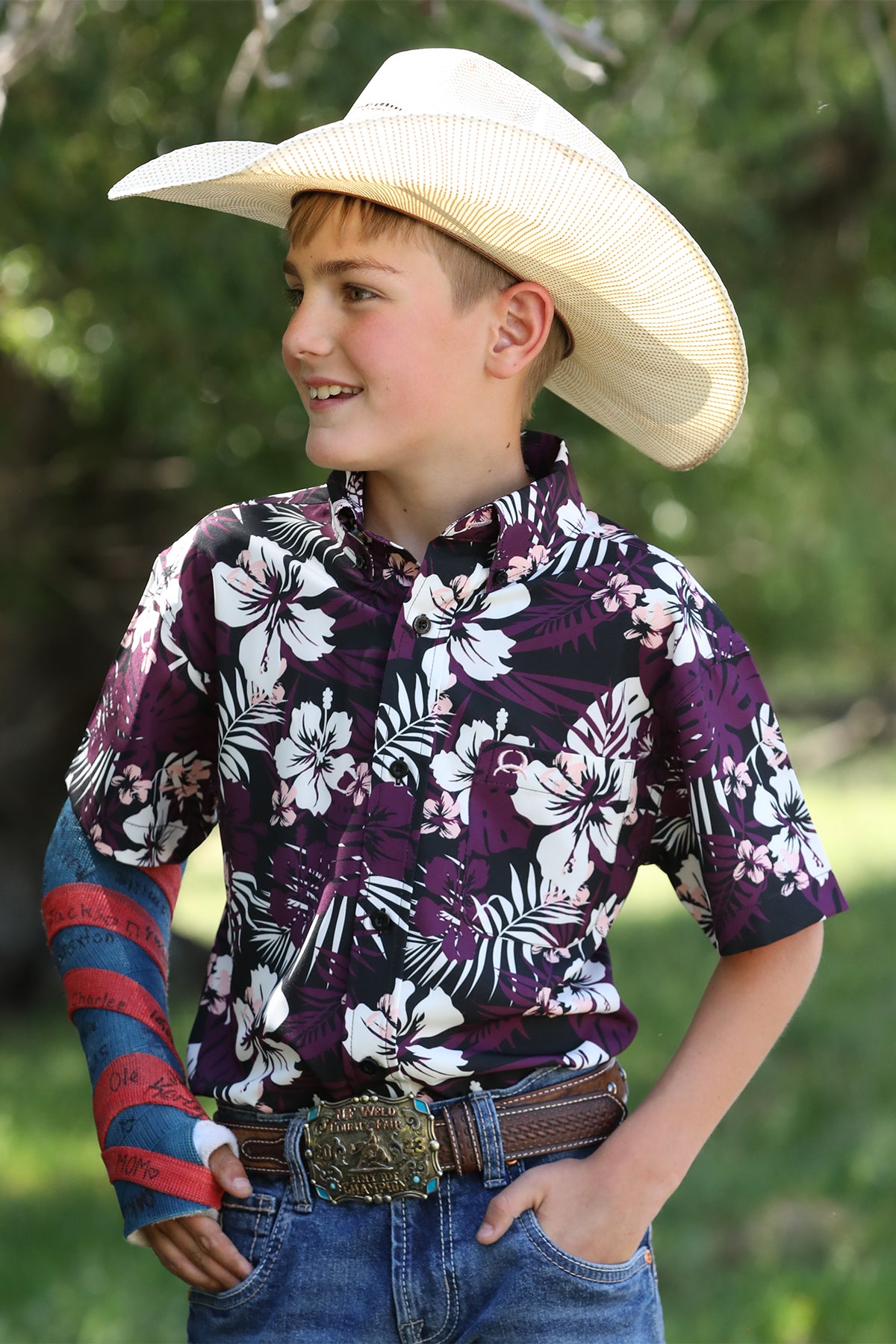 Boy standing outside wearing a patterned short sleeve button-down shirt with jeans, a belt, and a cowboy hat and has his arm in a cast