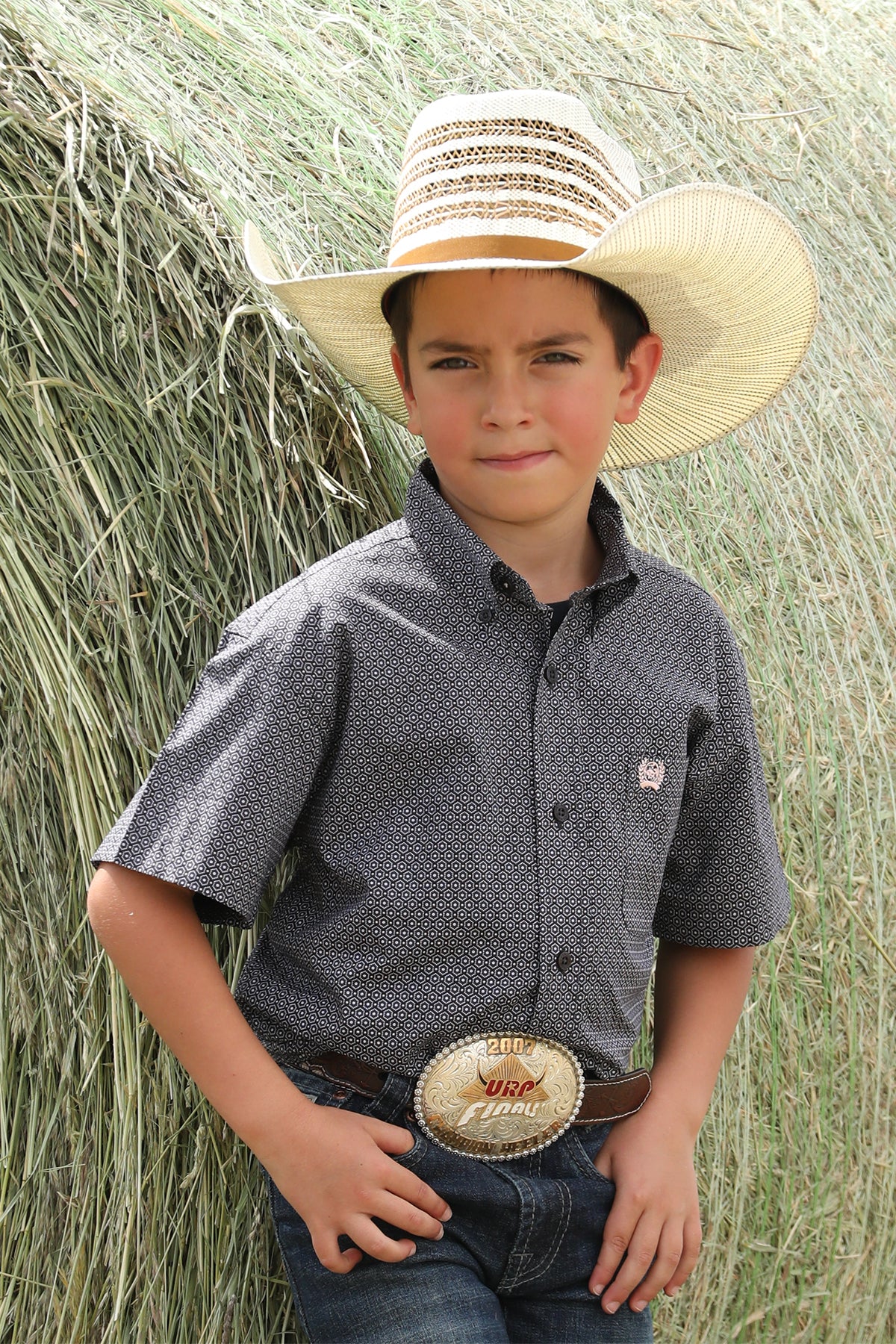 Boy standing outside in front of a bale of hay wearing a patterned short sleeve button-down shirt with jeans, a belt, and a cowboy hat