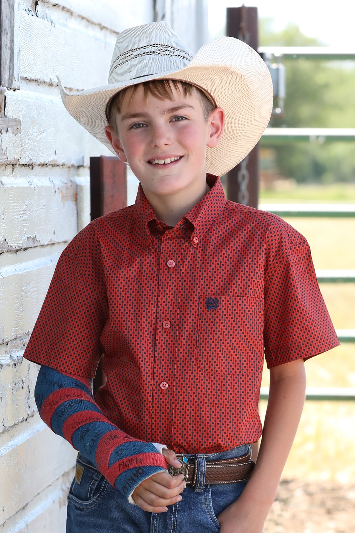 Boy standing outside wearing a red short sleeve button-down shirt with jeans, a belt, a cowboy hat and has his arm in a cast