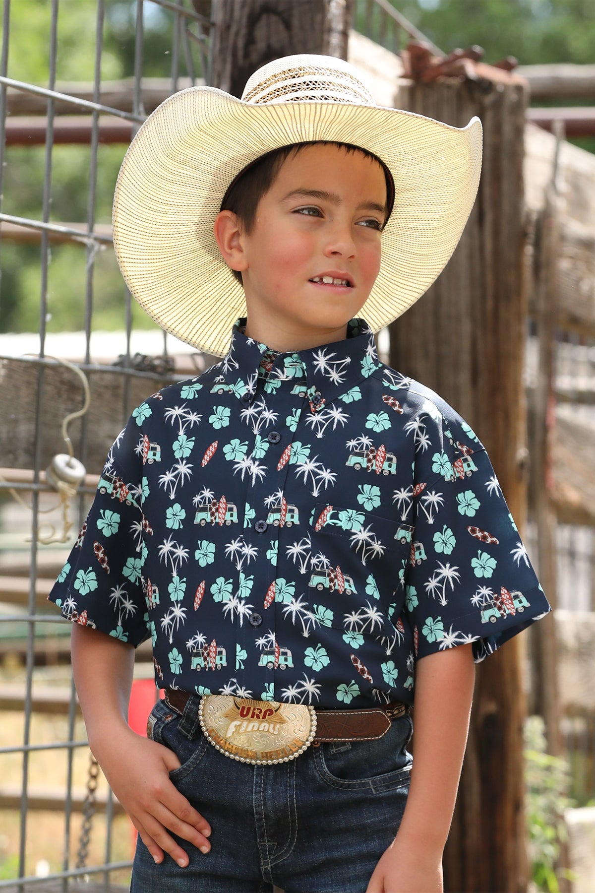 Boy standing outside in front of a fence wearing a patterned short sleeve button-down shirt with jeans, a belt, and a cowboy hat