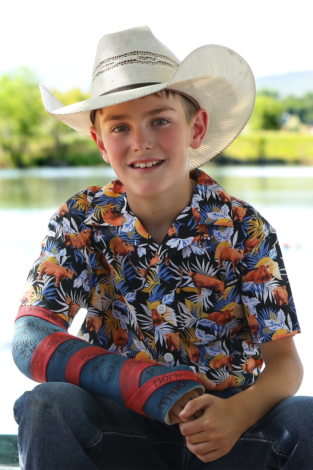 Boy sitting outside wearing a patterned short sleeve button-down with jeans and a cowboy hat with a cast on his arm
