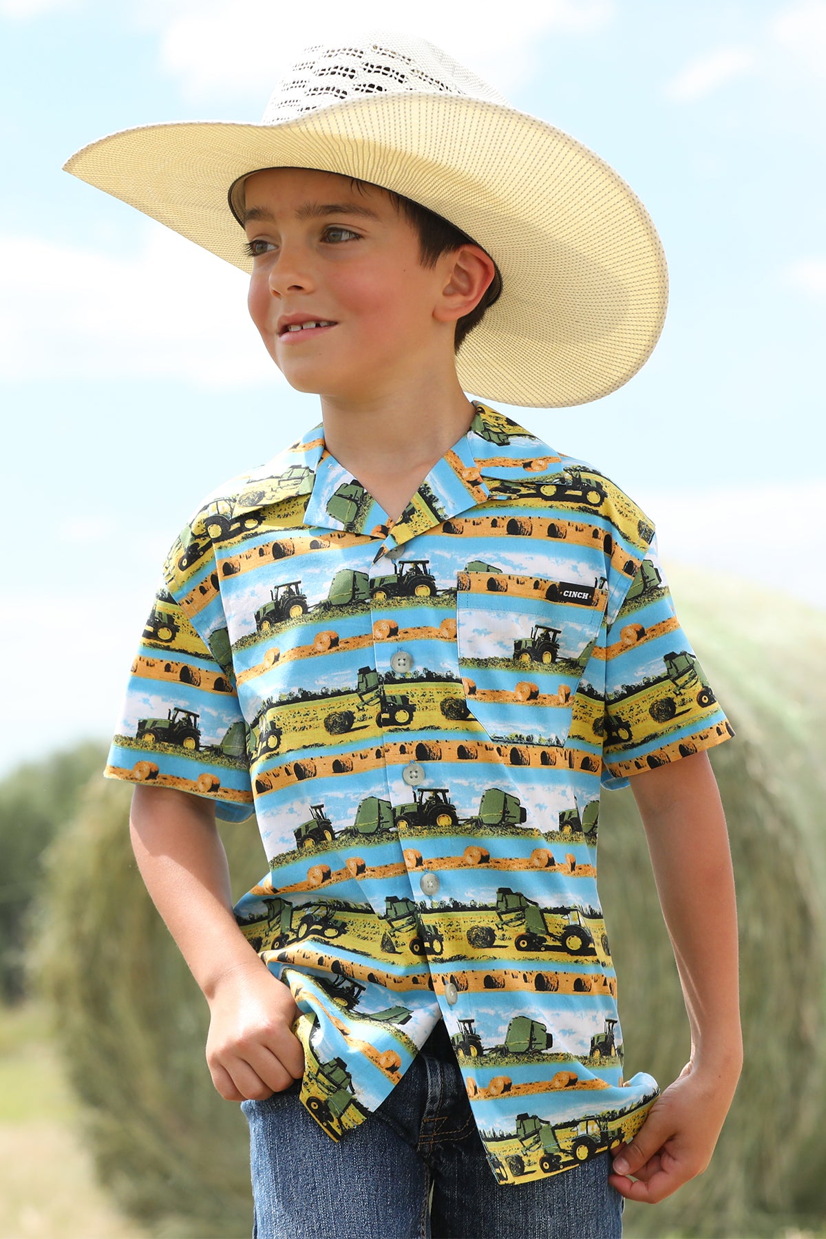 Boy standing outside in front of a bale of hay wearing a patterned short sleeve button-down shirt with jeans and a cowboy hat