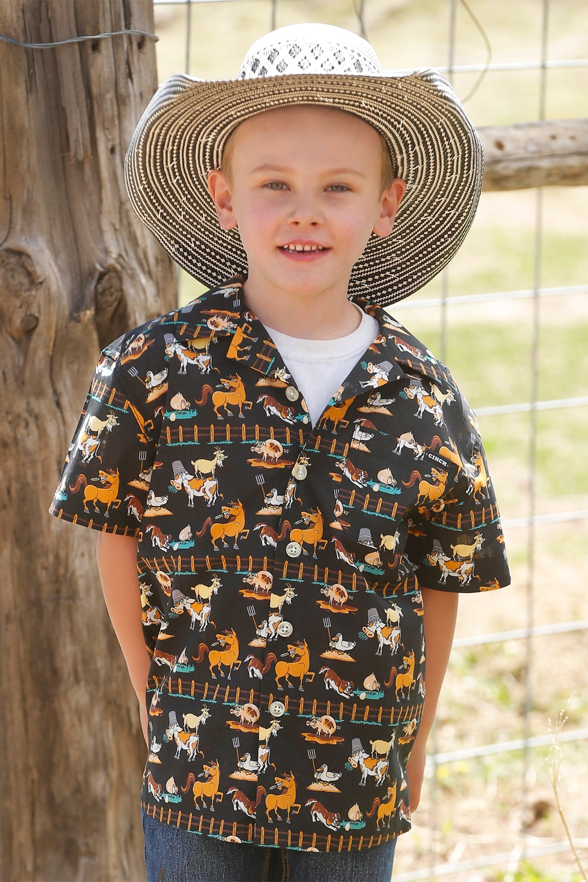 Little boy standing outside in front of a fence wearing a patterned short sleeve button-down shirt with jeans and a cowboy hat
