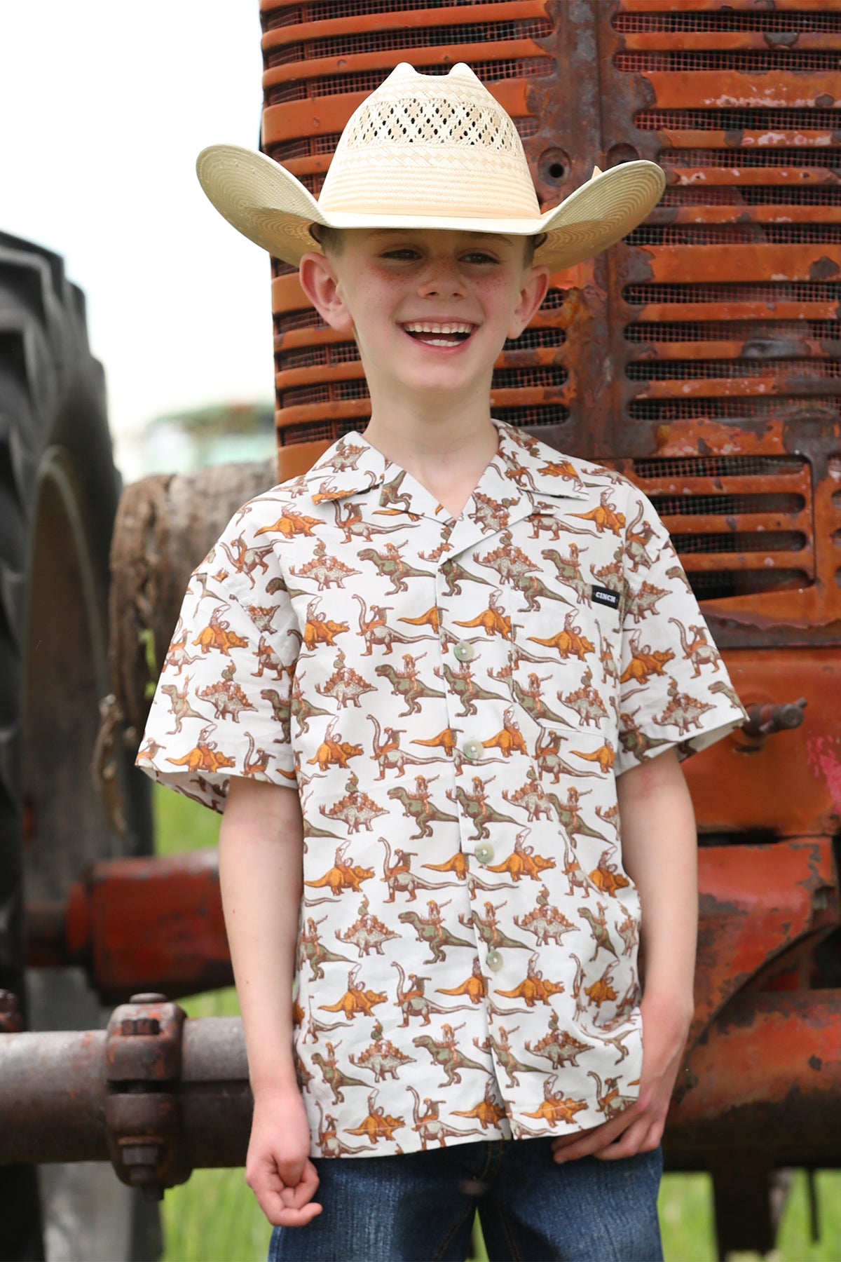 Boy standing outside in front of a tractor, wearing a patterned short sleeve button-down shirt with jeans and a cowboy hat
