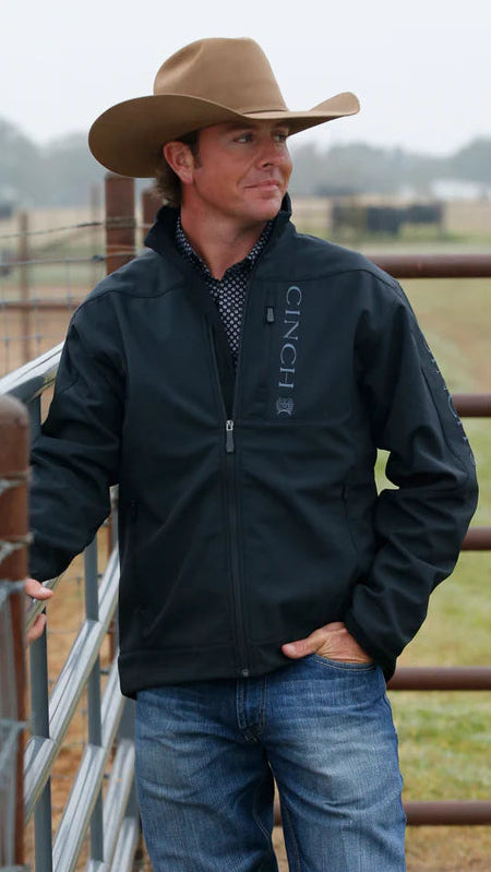 Man wearing a black jacket with 'Cinch' branding and a cowboy hat, standing in a rural setting with horses and a fence.