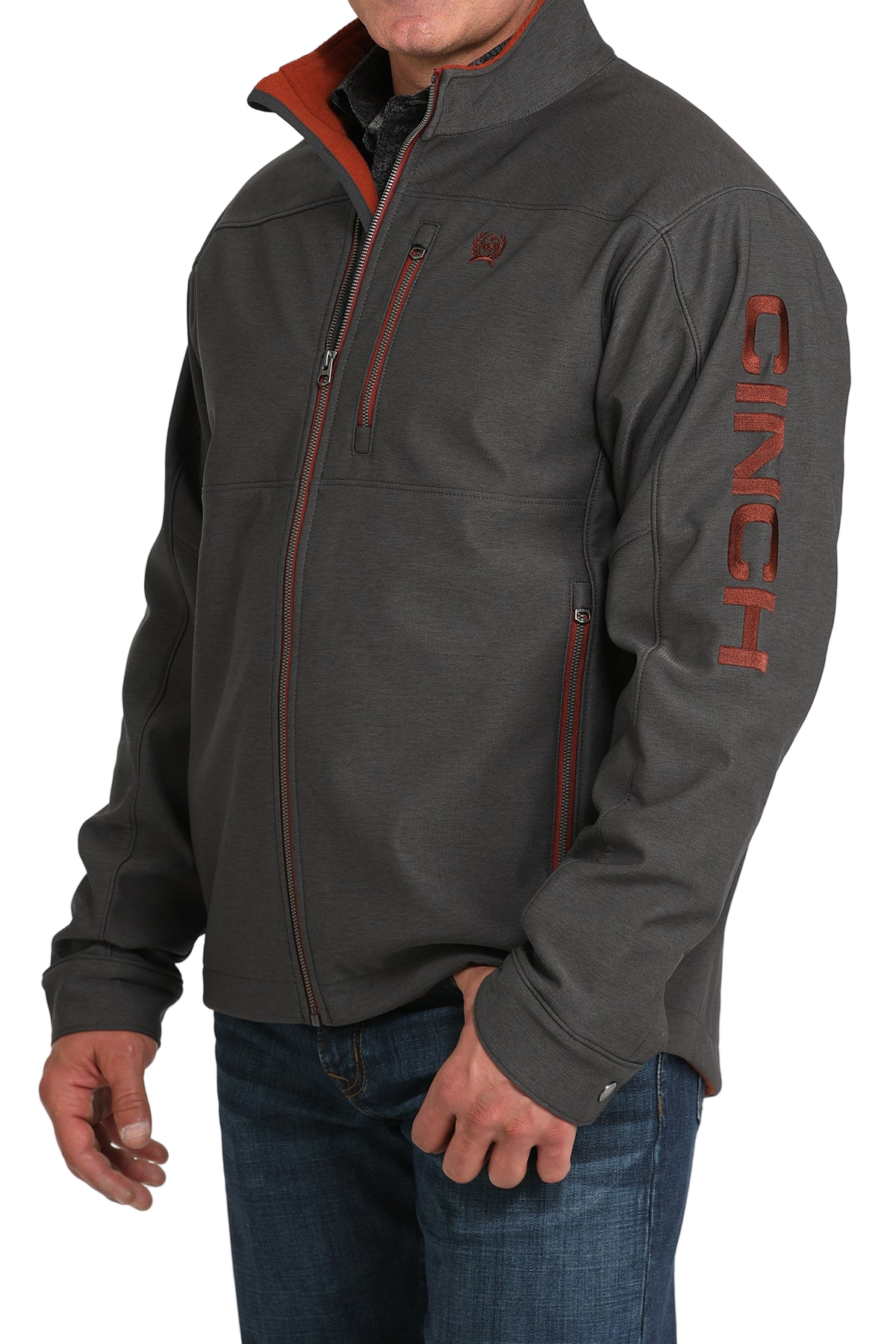 Man wearing a charcoal gray softshell jacket with bold red accents and “CINCH” lettering down the sleeve, paired with blue jeans