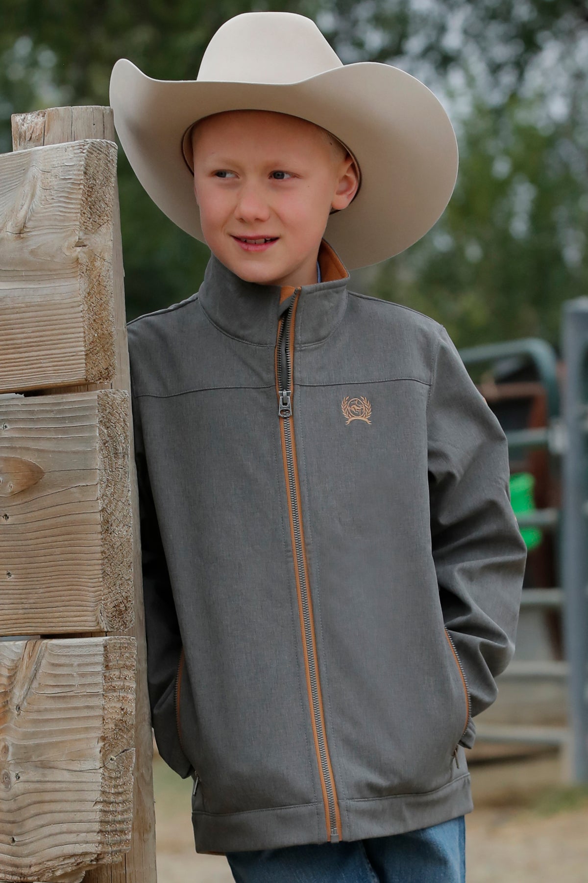 Boy standing outside leaning against a wood fence, wearing a 'CINCH' jacket, jeans and a cowboy hat