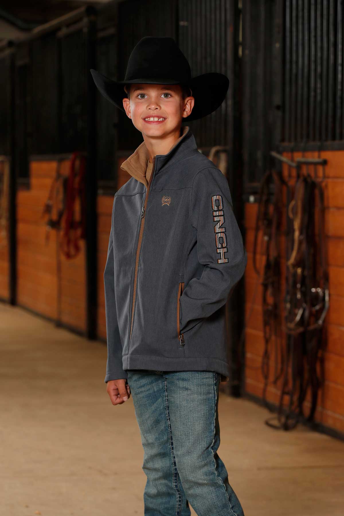 A young boy wearing a black cowboy hat, gray jacket, and jeans stands smiling in a barn hallway with tack and wooden stalls in the background