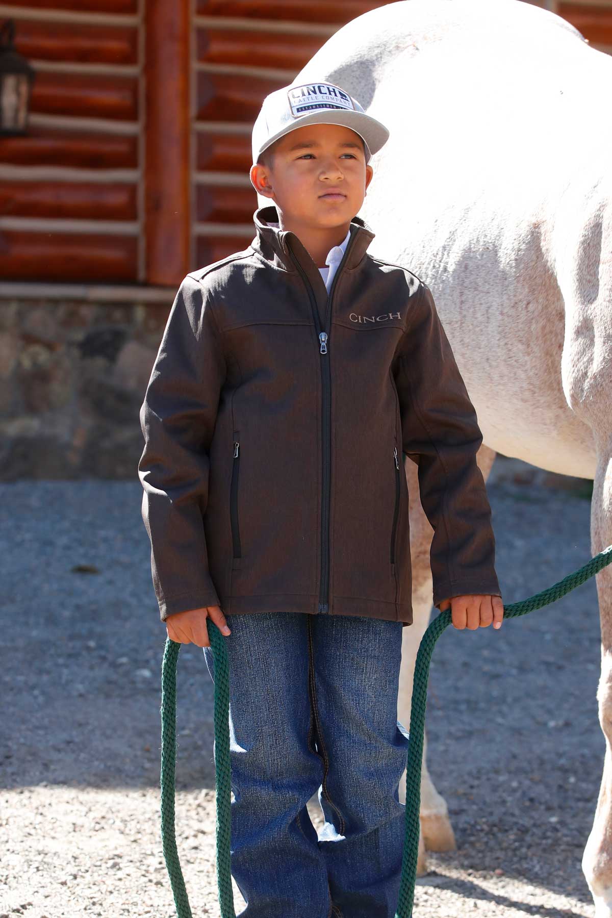 A young boy wearing a gray cap, dark jacket, and jeans stands outside holding a green lead rope next to a white horse