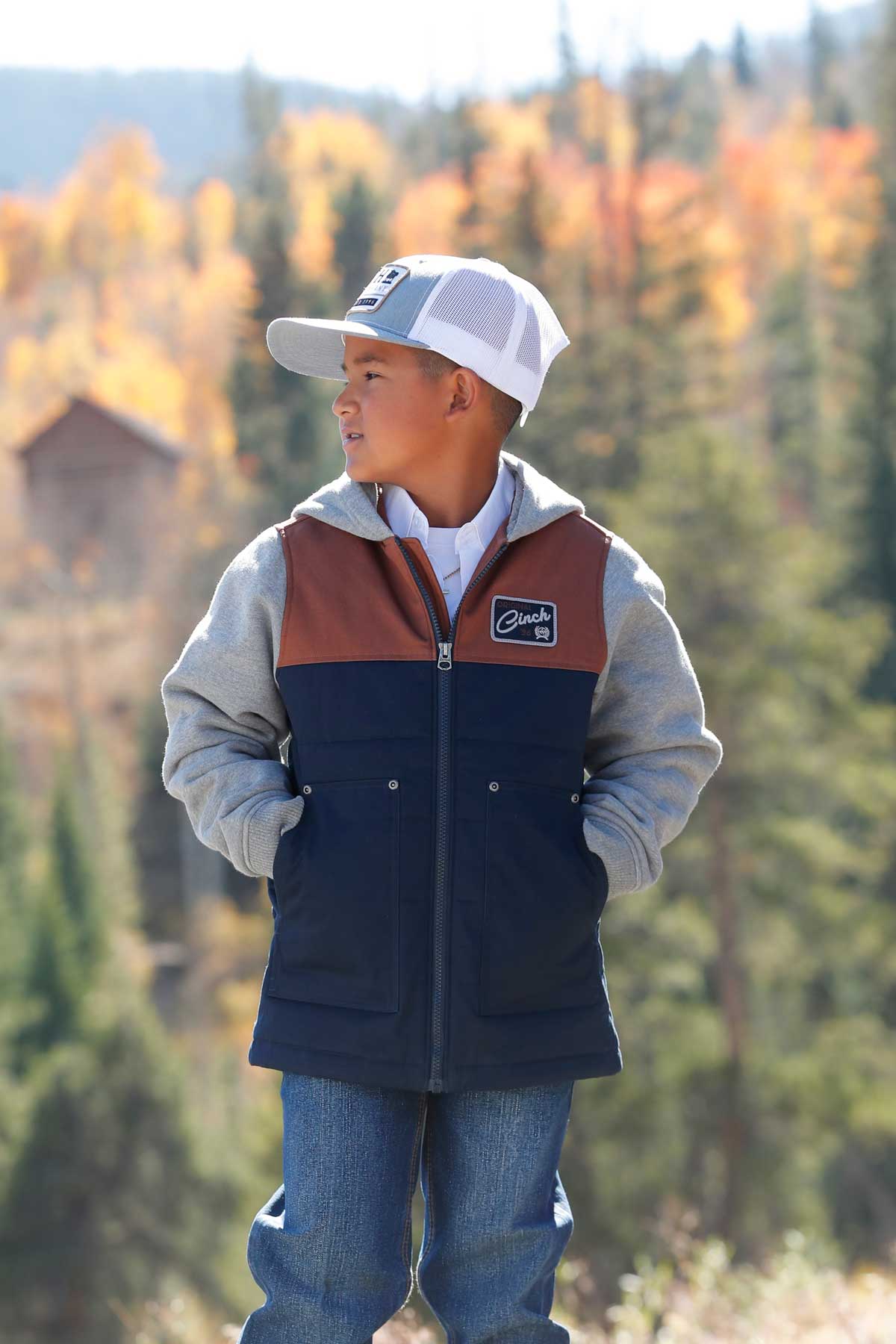 A young boy wearing a gray cap, color-block jacket, and jeans stands outdoors with autumn trees in the background