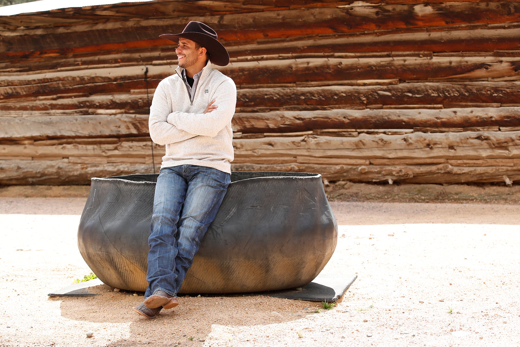 Man wearing a khaki 1/4 zip sweater with jeans and a cowboy hat, leaning on a metal structure outside