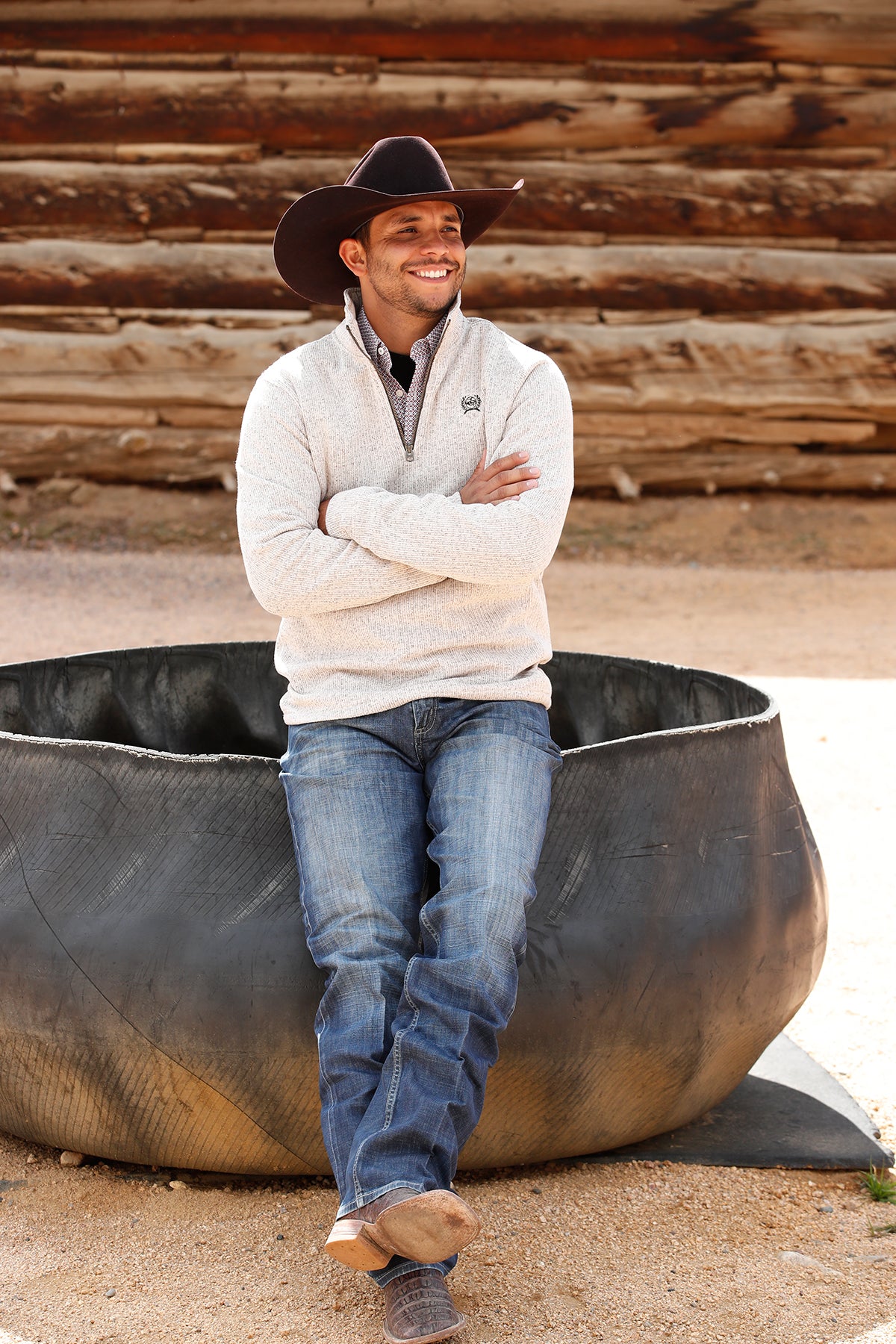 Man wearing a khaki 1/4 zip sweater with jeans and a cowboy hat, leaning on a metal structure outside