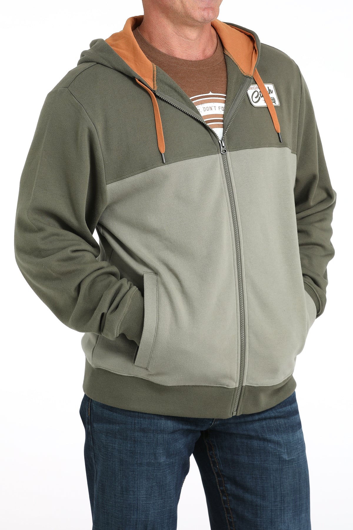 Man wearing a two-tone green zip-up hoodie with an orange hood lining and blue jeans displayed on a white background.