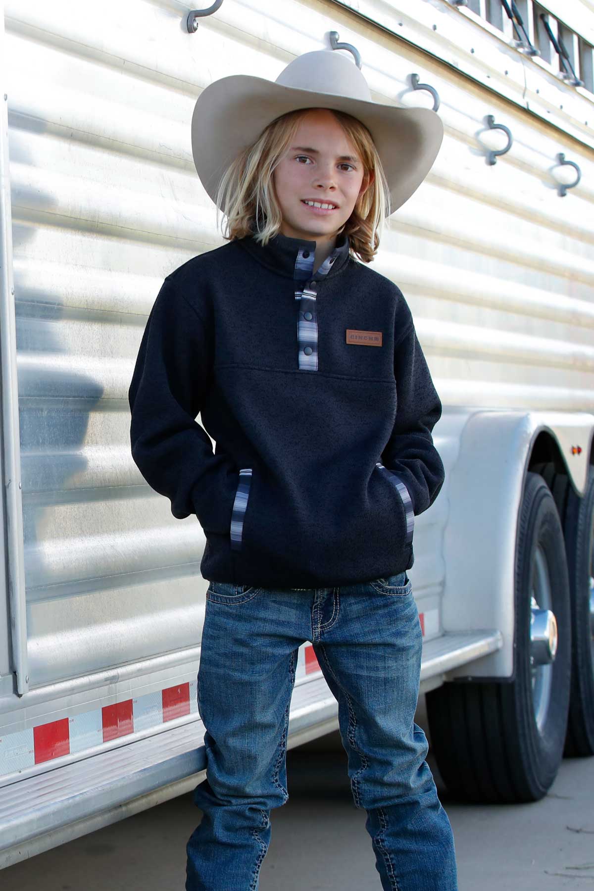 A young boy wearing a white cowboy hat, dark pullover, and jeans stands in front of a silver horse trailer with hands in his pockets