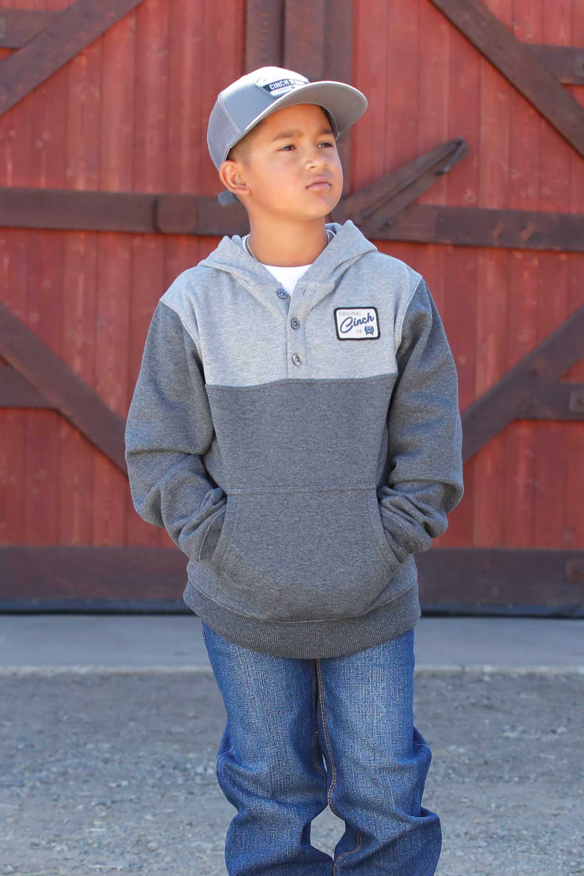 Child wearing a gray hoodie with a logo and blue jeans standing in front of a red barn door.