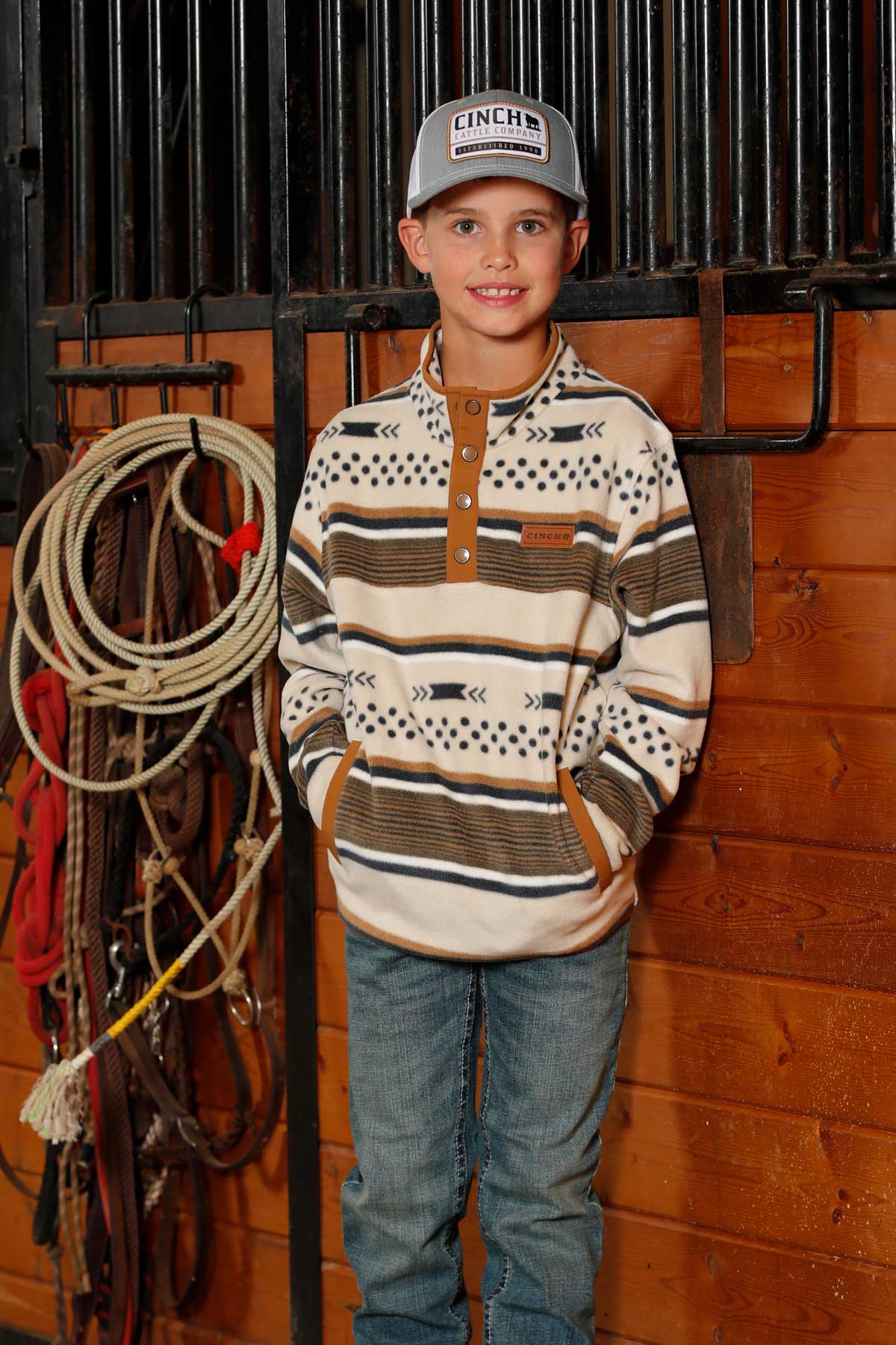A young boy wearing a gray cap, patterned pullover, and jeans stands in a barn with ropes and tack hanging on the wall behind him