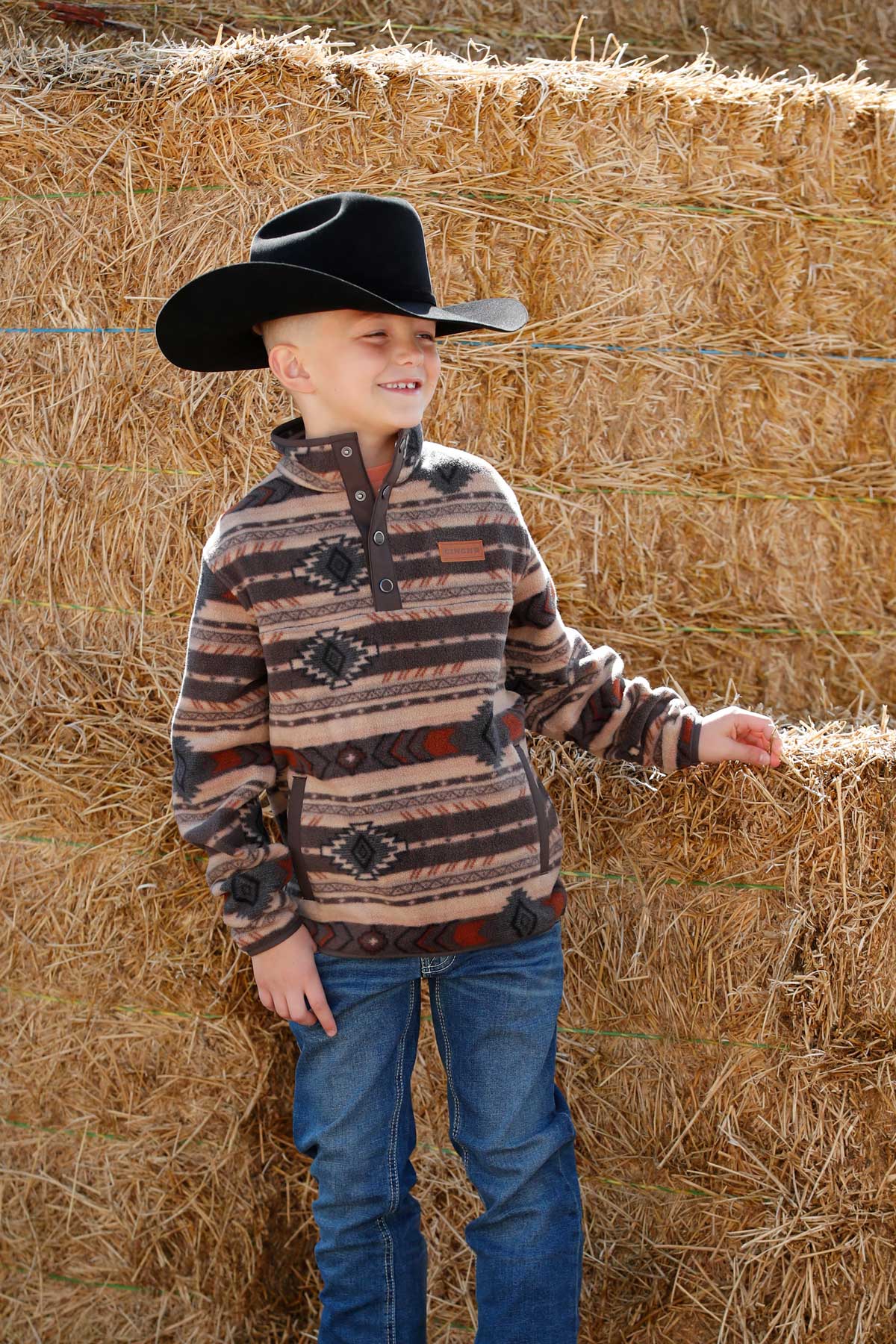 A young boy wearing a black cowboy hat, patterned pullover, and jeans stands in front of stacked hay bales, smiling