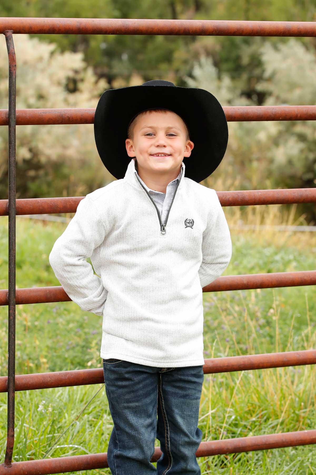A young boy wearing a black cowboy hat, light pullover, and jeans stands in front of a metal fence, smiling