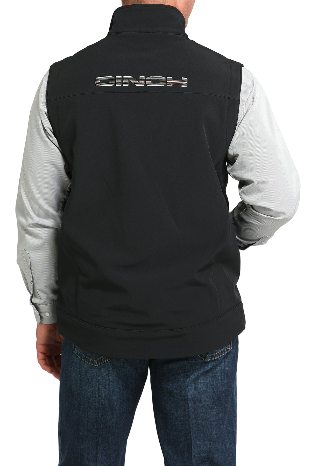 A man shown from the back wearing a black vest with “CINCH” embroidered across the upper back, layered over a light gray long-sleeve shirt and paired with blue jeans