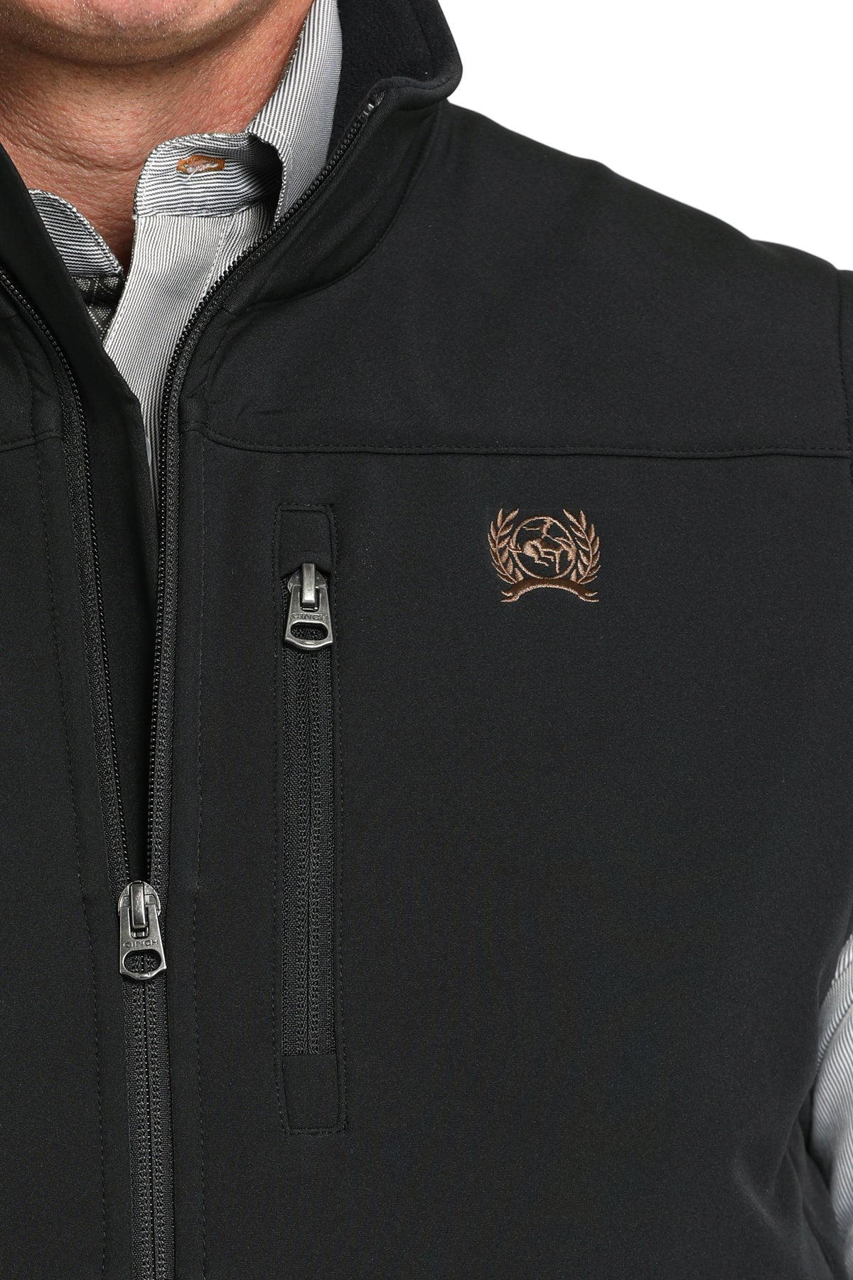 A close-up of a black men’s vest showing the front zipper, a vertical chest pocket with a zipper, and a small embroidered logo on the left chest. A gray collared shirt is visible underneath