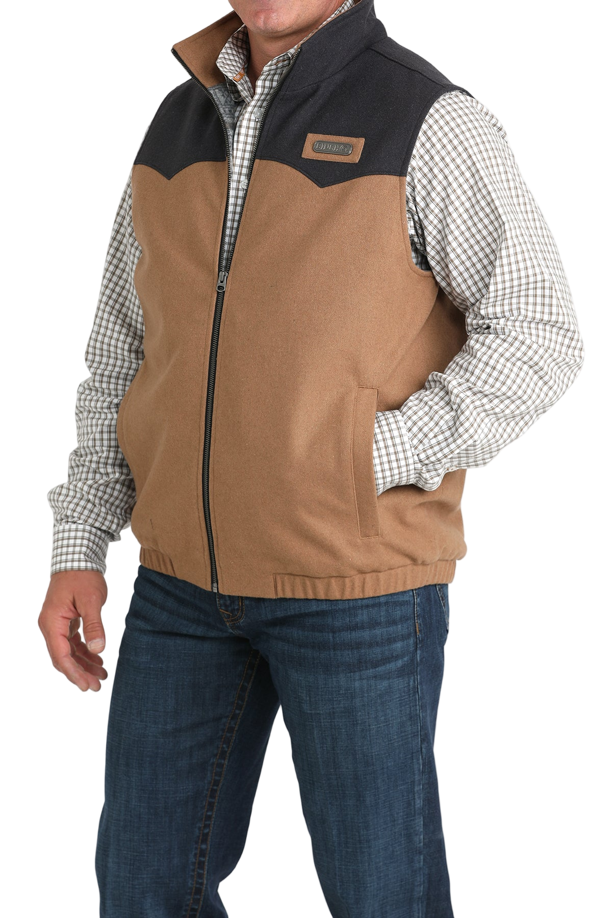 A man wearing a two-tone zip-up vest with a tan body and dark charcoal upper panels over a plaid button-down shirt and jeans