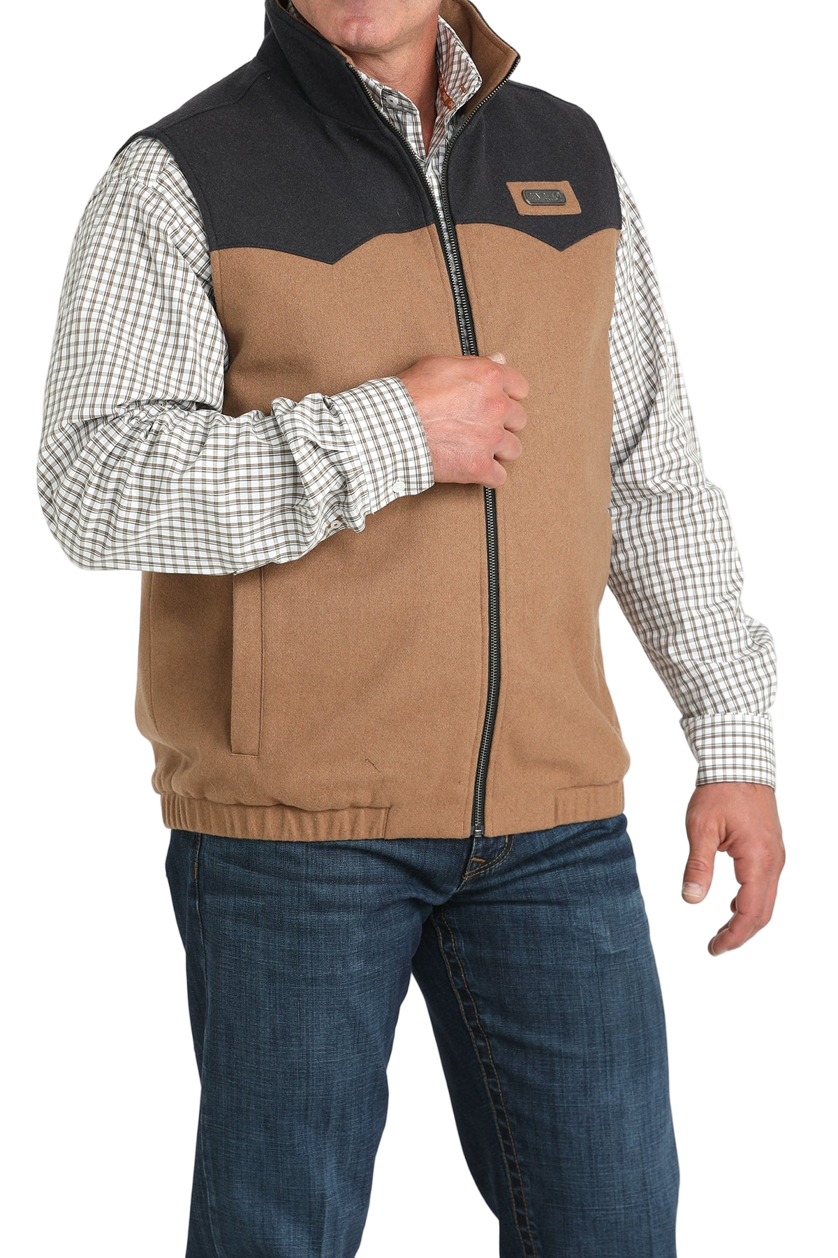 A man wearing a two-tone zip-up vest with a tan body and dark charcoal upper panels over a plaid button-down shirt and jeans