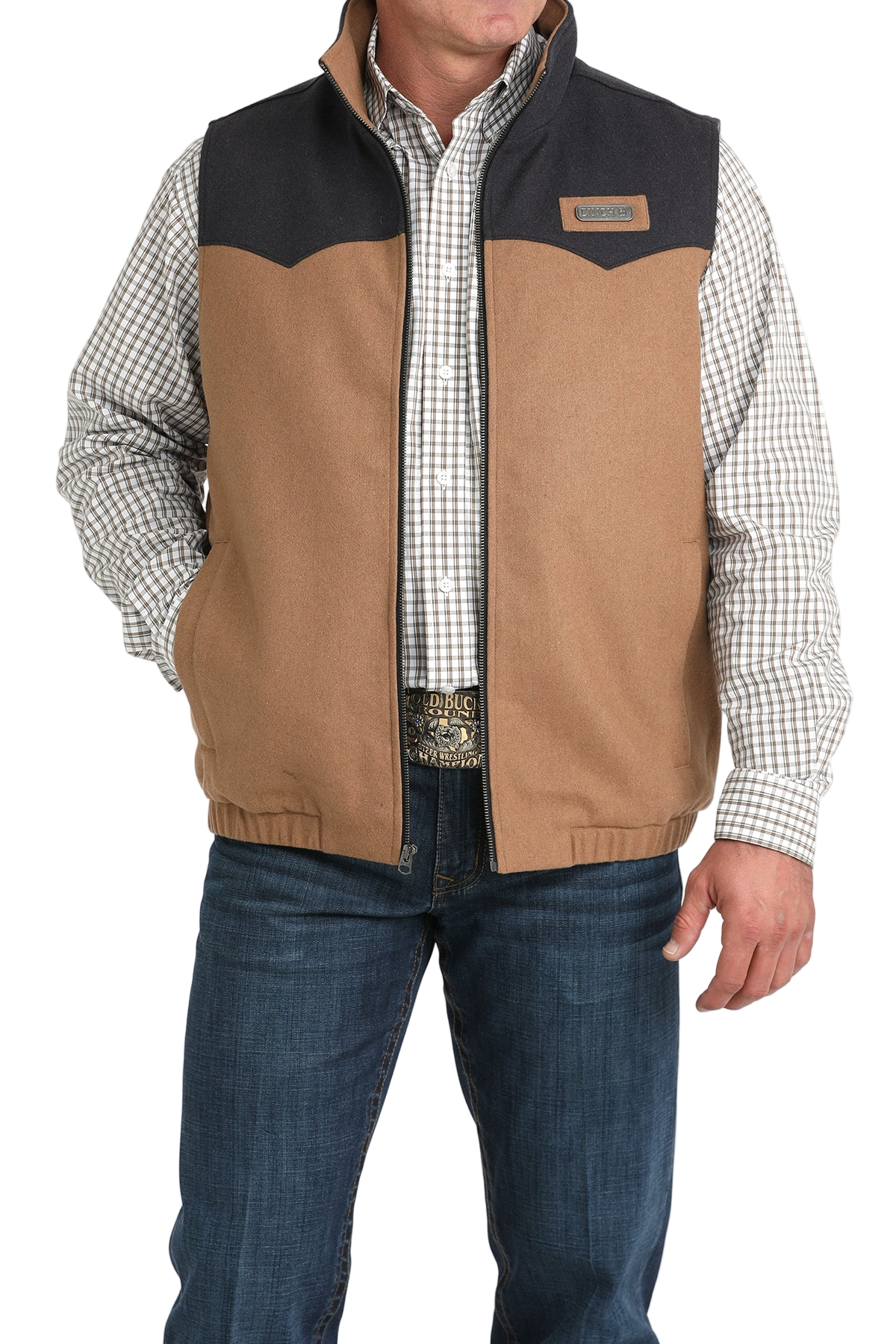 A man wearing a two-tone zip-up vest with a tan body and dark charcoal upper panels over a plaid button-down shirt and jeans