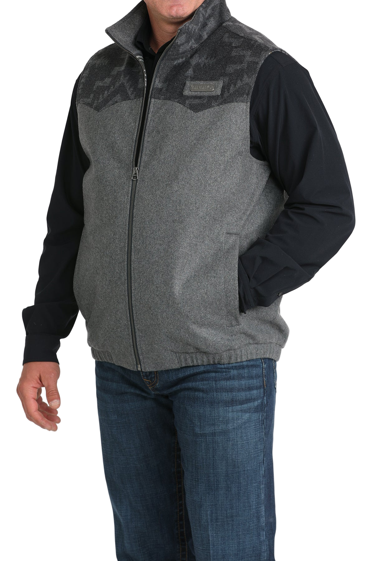 A man wearing a grey zip-up vest with a darker patterned yoke, paired with a black long-sleeve shirt and blue jeans, standing with one hand in his pocket