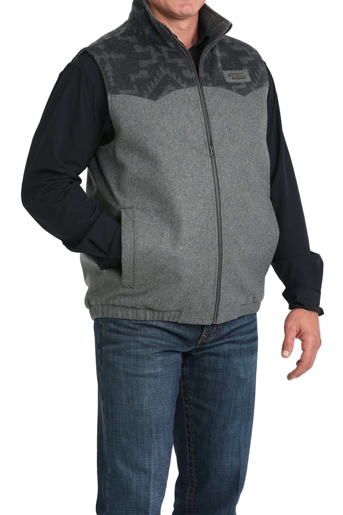 A man wearing a grey zip-up vest with a darker patterned yoke, paired with a black long-sleeve shirt and blue jeans, standing with one hand in his pocket