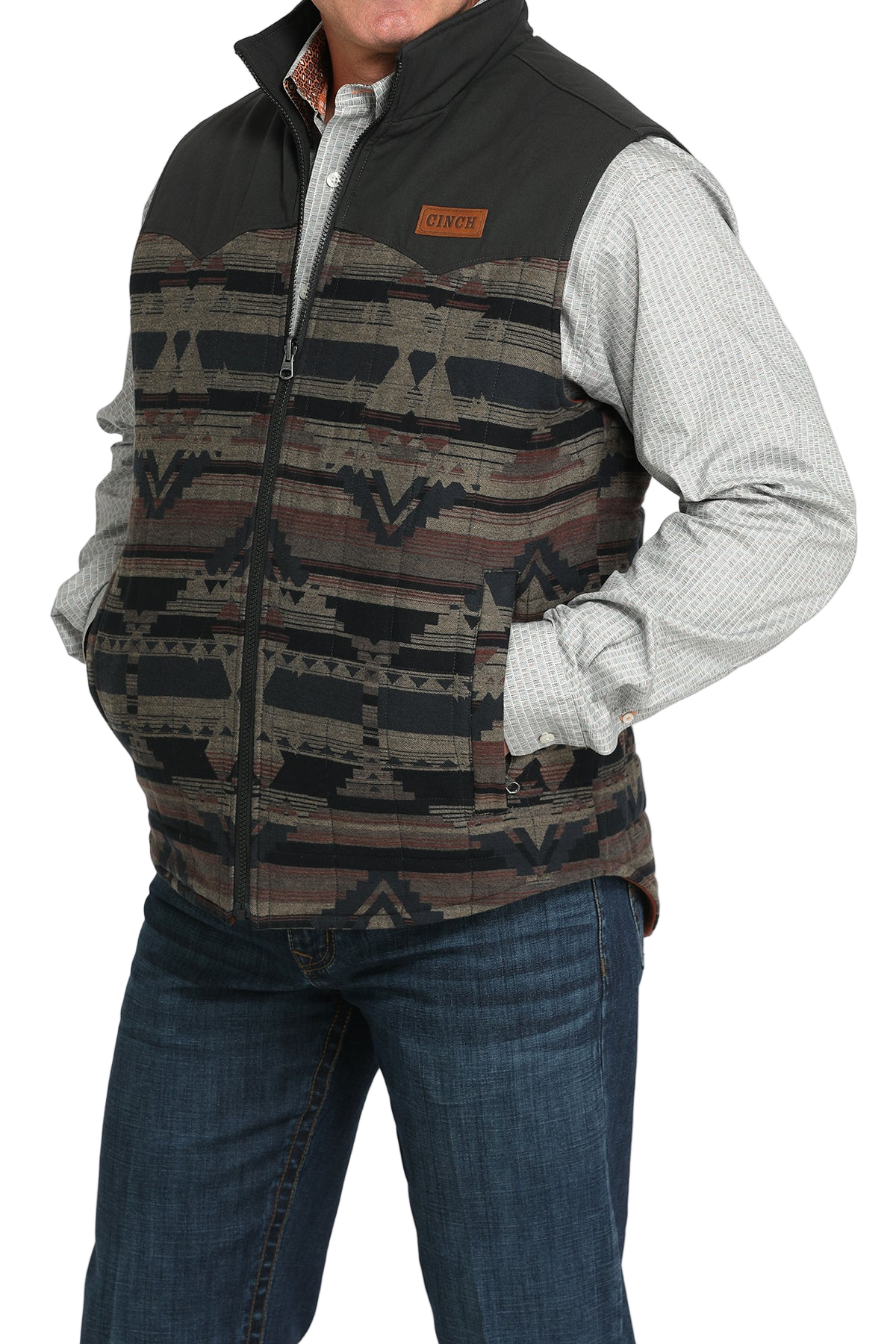 A man wears a patterned zip-front vest with black shoulders over a light gray checkered shirt and jeans