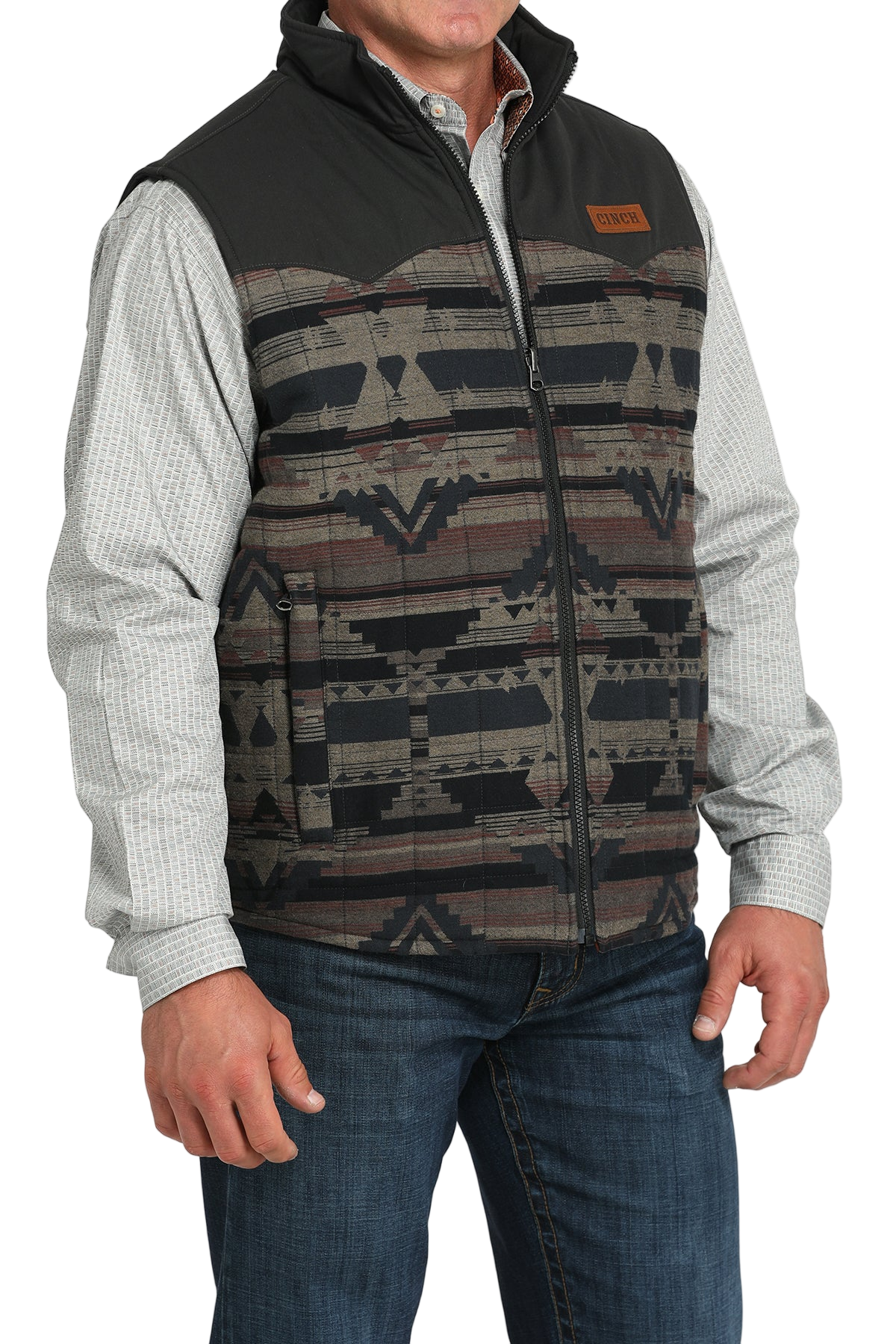A man wears a patterned zip-front vest with black shoulders over a light gray checkered shirt and jeans