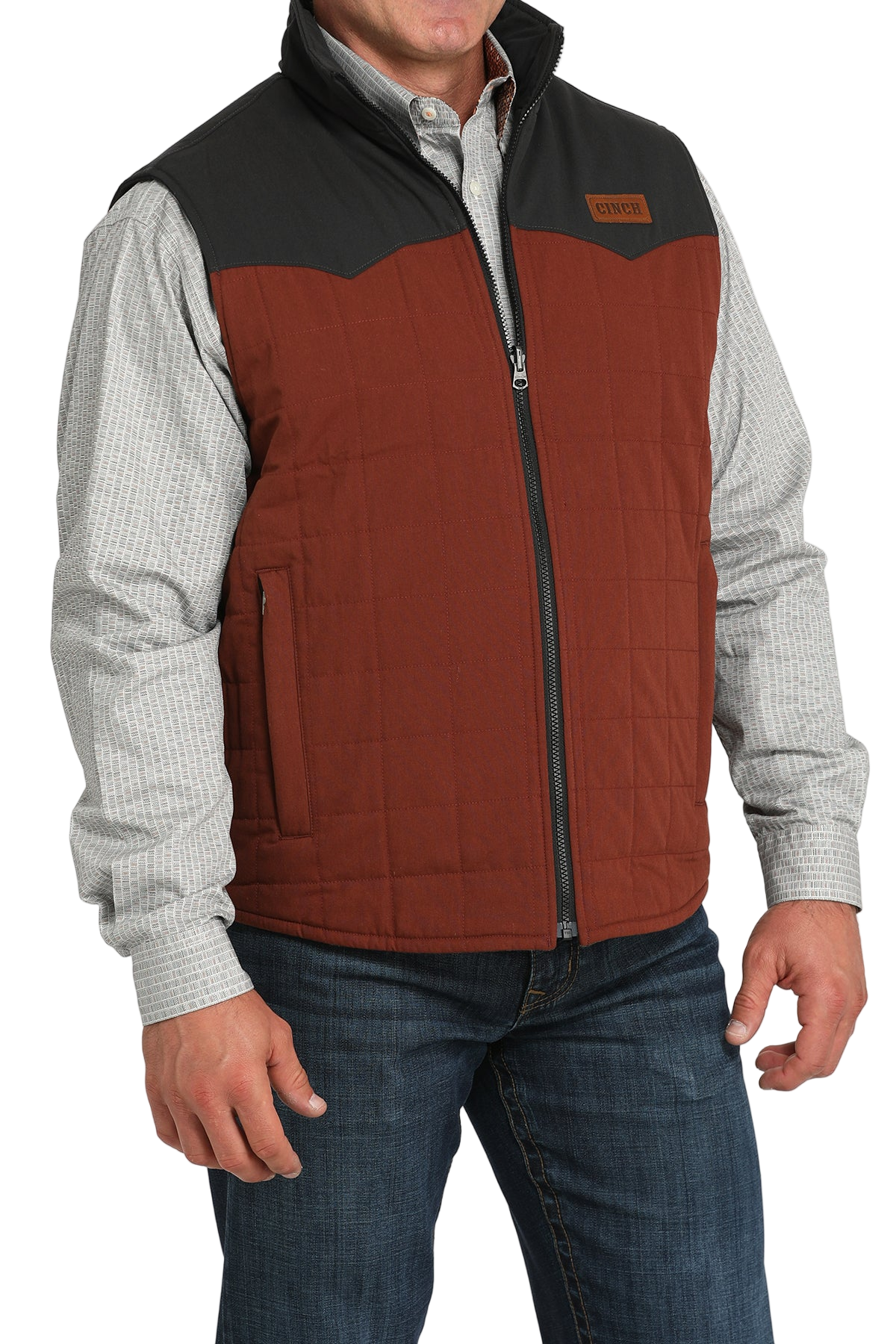 A man wears a two-tone orange and brown zip-front vest over a light gray checkered shirt and jeans