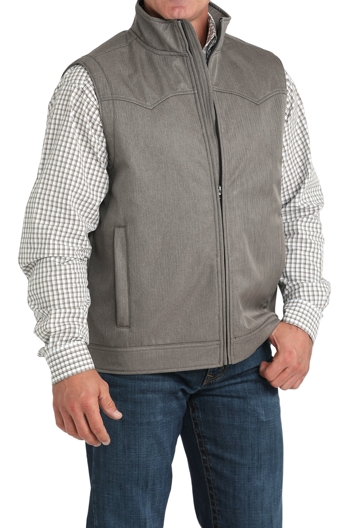 A man wearing a light gray zip-up vest over a white and brown checkered button-down shirt, paired with blue jeans