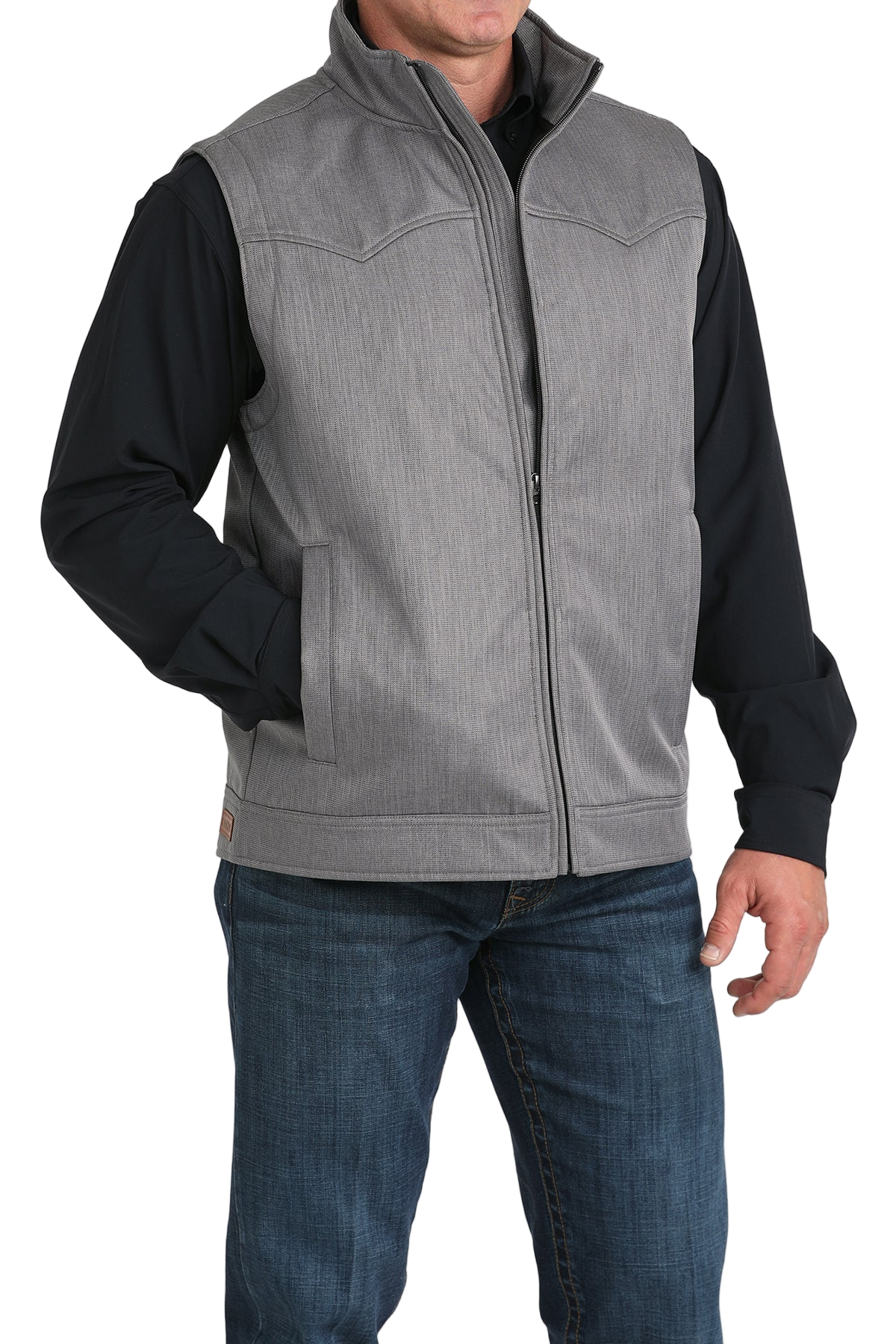 A man wears a light gray zip-front vest over a black long-sleeve shirt and blue jeans