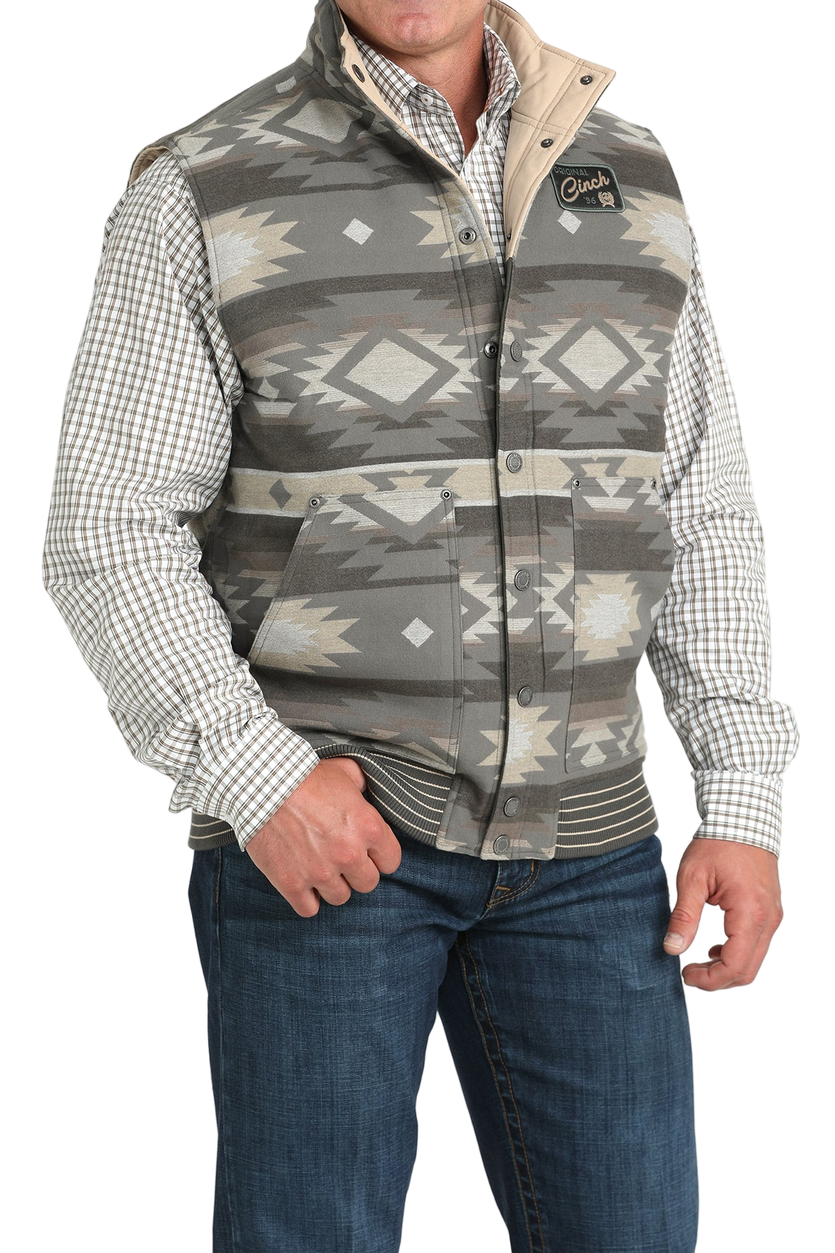 A man wearing a gray and beige Aztec-patterned vest over a checkered button-down shirt, paired with blue jeans